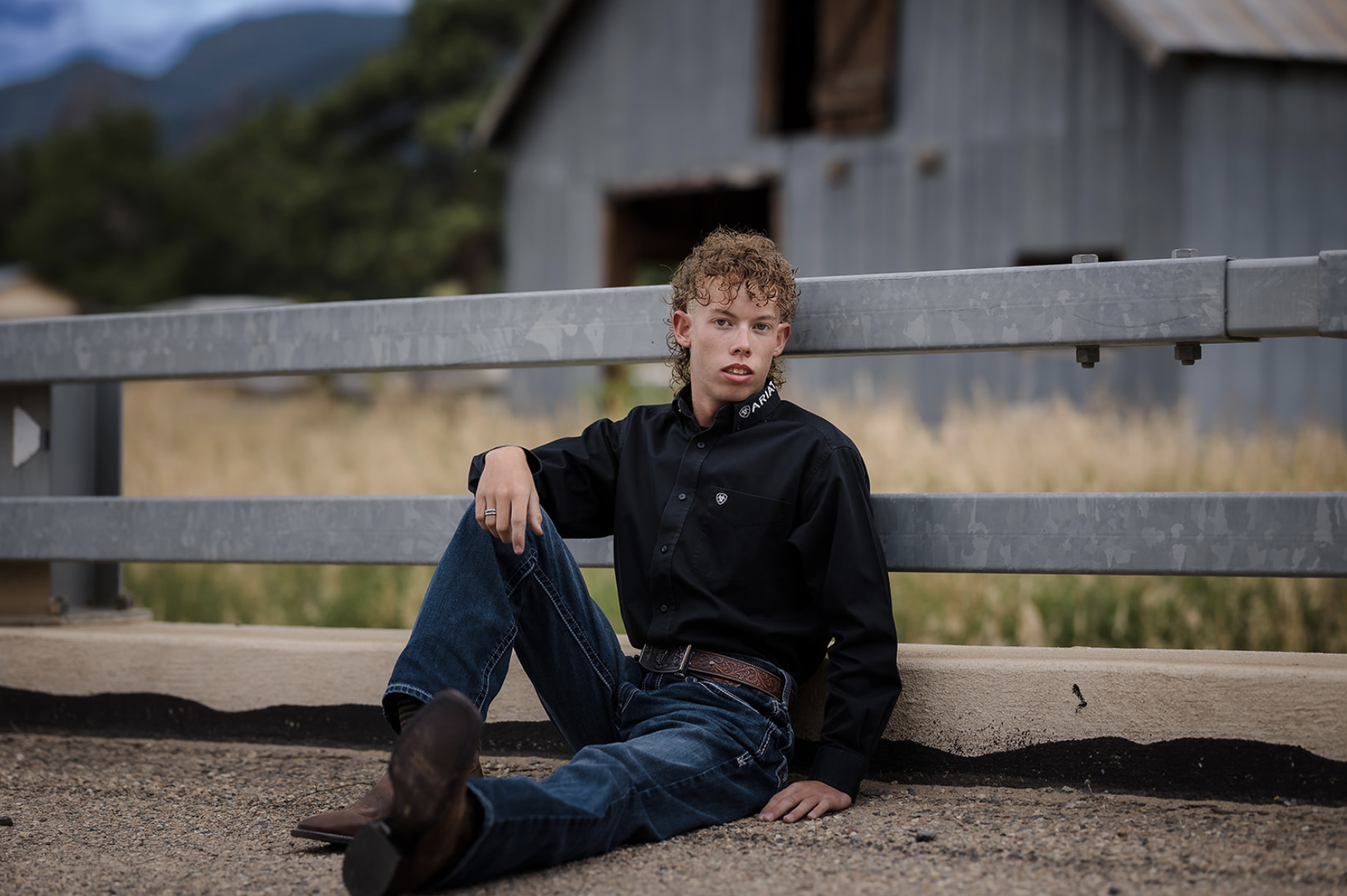 A young man with curly hair sitting on the ground against a metal railing in front of a rustic barn and mountains, wearing a black button-up shirt and jeans.