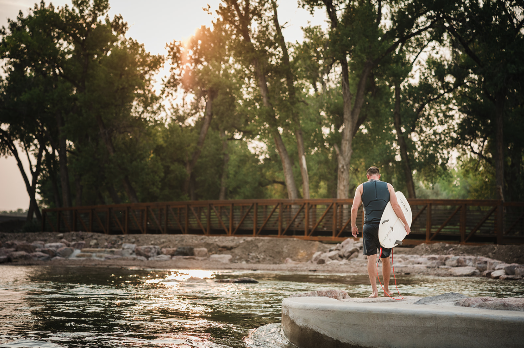 A man walking on a concrete slab towards the water holding a surfboard under his arm, with a forest and sunset in the background.
