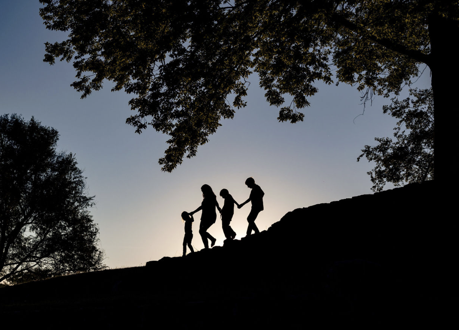 Young man sitting outdoors on grass near a tree, wearing a colorful floral shirt and blue jeans, with a thoughtful expression.