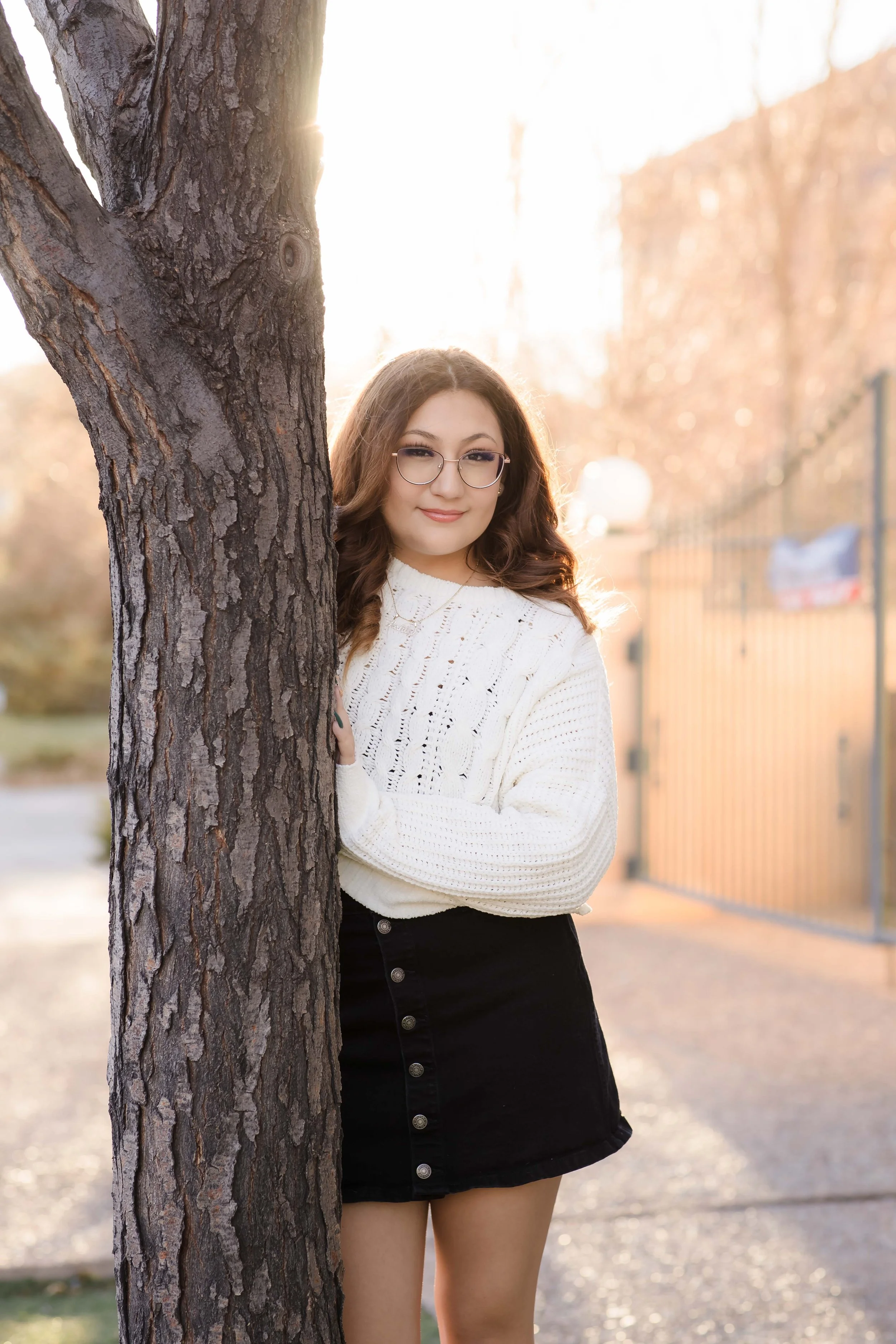 A young woman with glasses and brown hair peeking from behind a tree, outdoors in a park or street during golden hour, with sunlight behind her and a fence in the background.