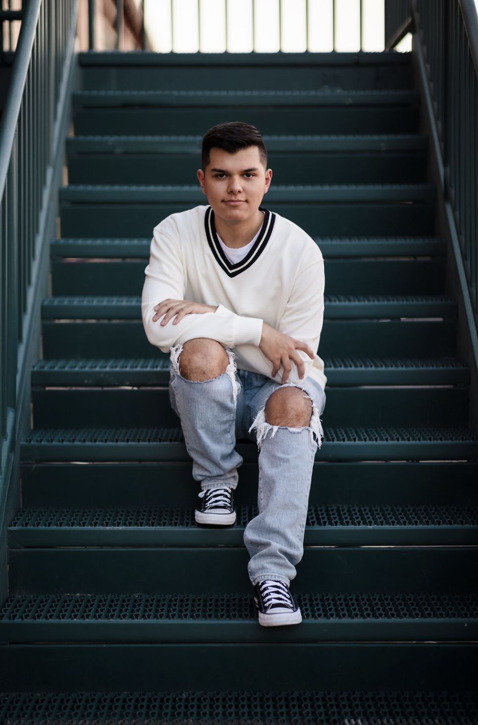 A young man sitting on outdoor metal stairs, wearing a white sweater with black trim, ripped jeans, and sneakers, looking at the camera.