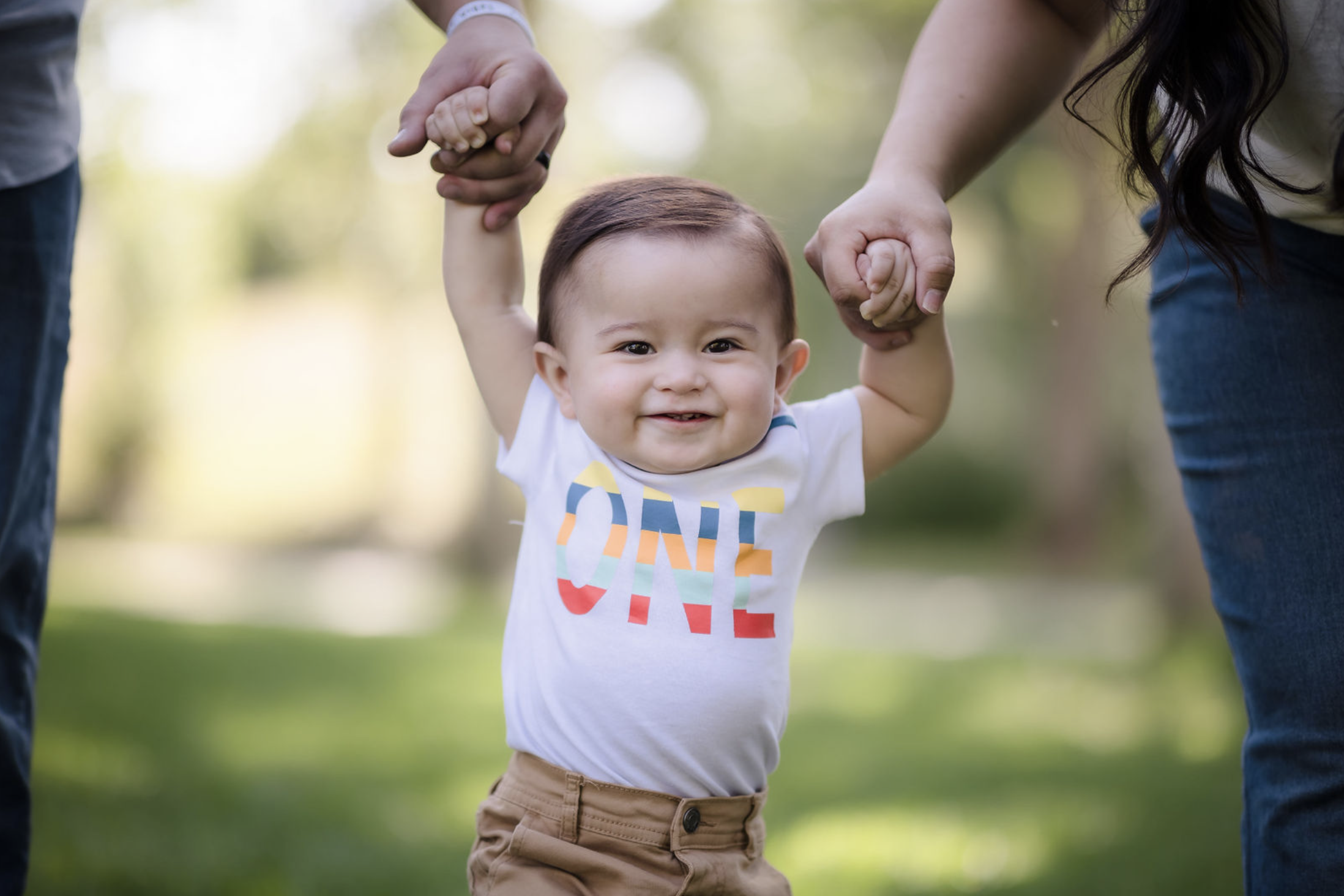 Young man sitting outdoors on grass near a tree, wearing a colorful floral shirt and blue jeans, with a thoughtful expression.