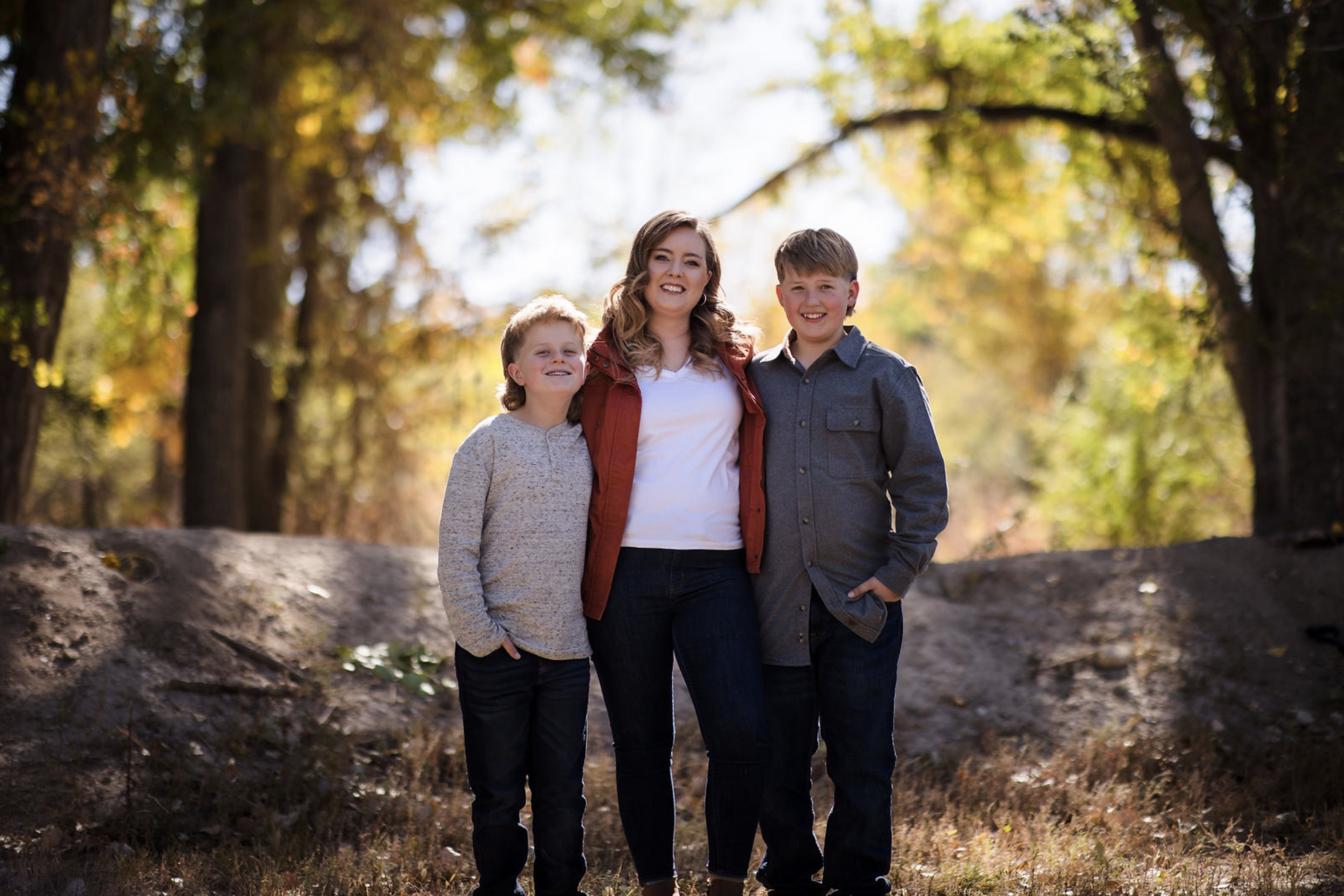 Young man sitting outdoors on grass near a tree, wearing a colorful floral shirt and blue jeans, with a thoughtful expression.