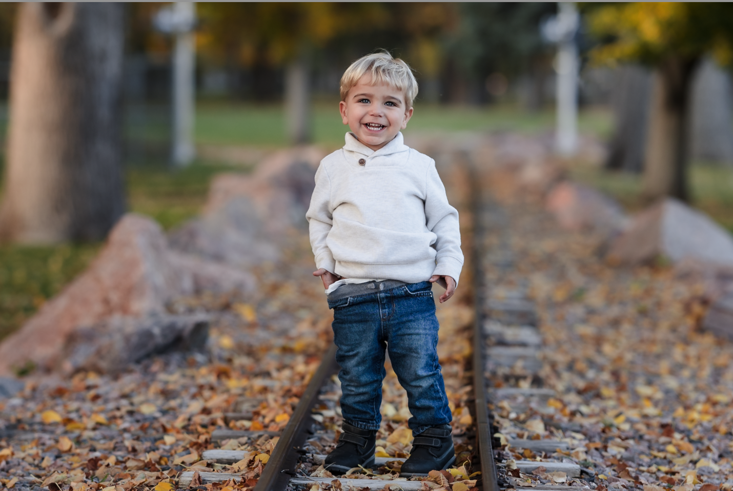 A young girl and boy hugging on a dirt path in a wooded area with autumn leaves, with a man and woman standing in the background, slightly out of focus.