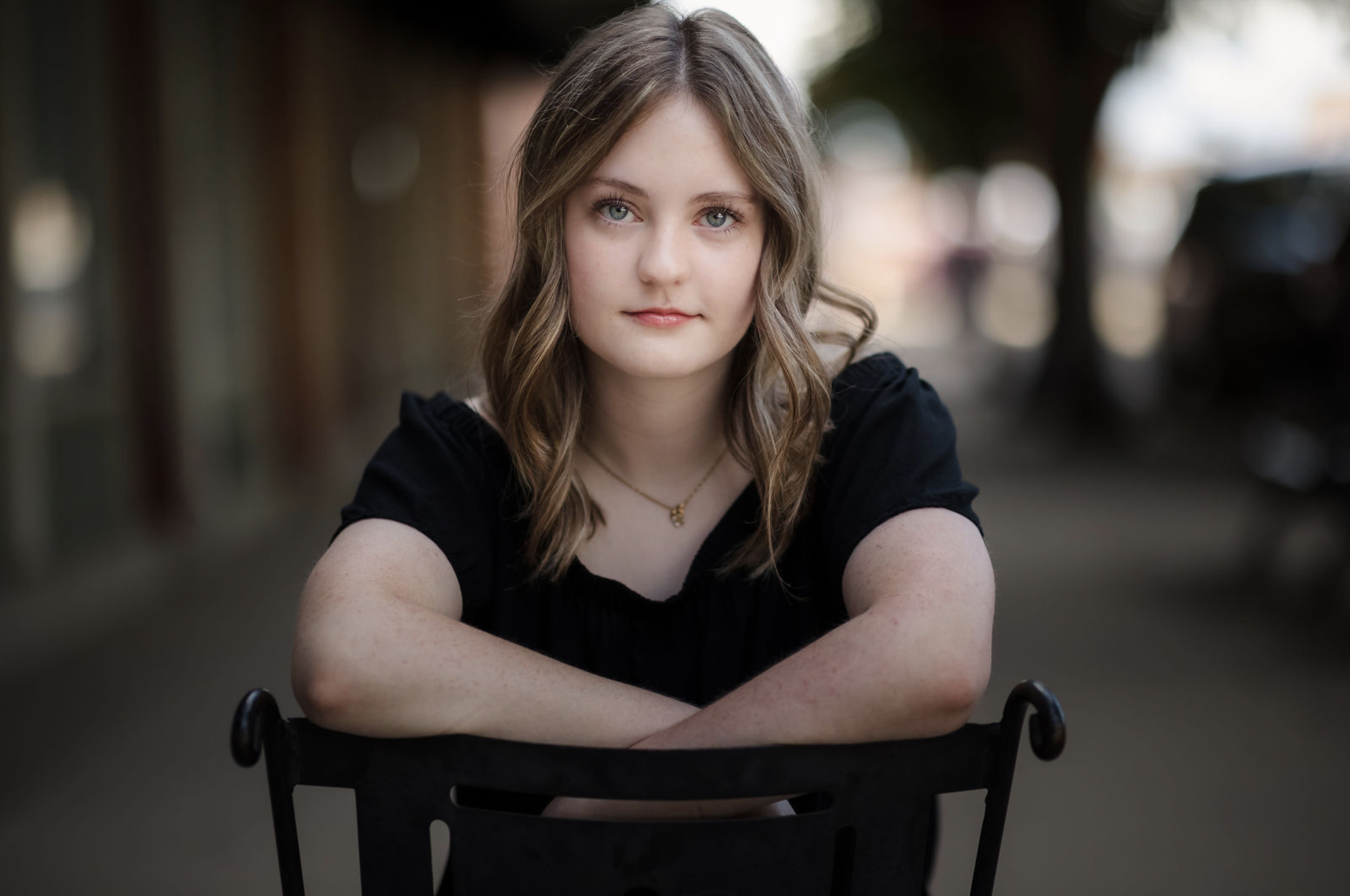 A young woman with wavy blonde hair, blue eyes, and light skin, wearing a black top and a gold necklace, sitting outdoors at a table with her arms crossed, looking at the camera with a neutral expression.