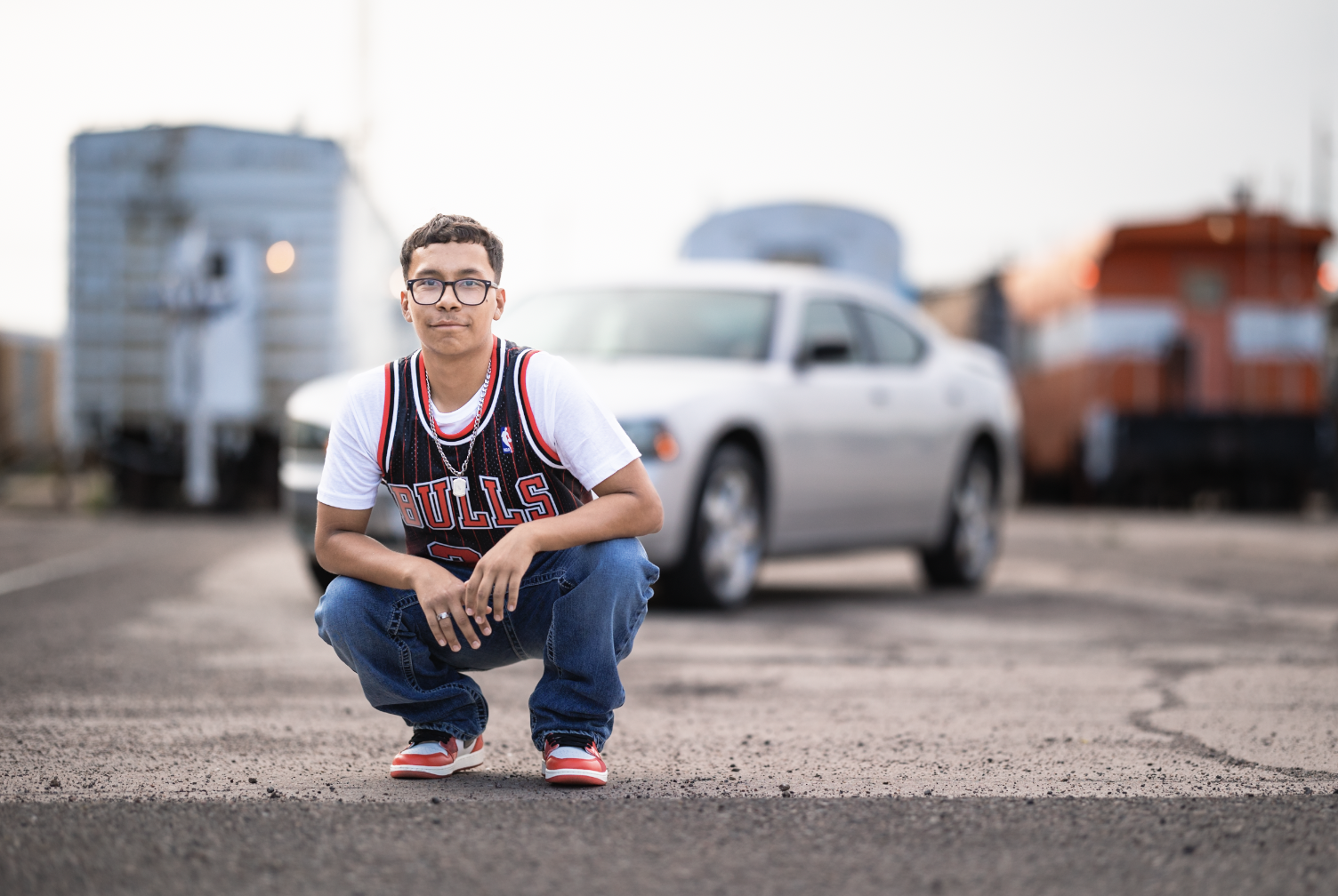 A young man wearing glasses, a Chicago Bulls basketball jersey, and jeans crouching on a rough pavement in front of a silver car, with train cars blurred in the background.