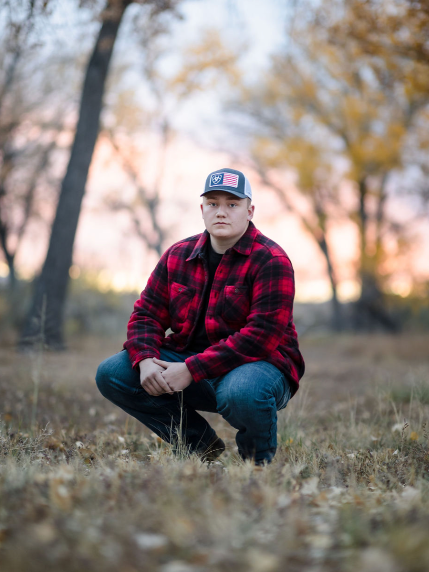 A young man crouching outdoors on a fall day, wearing a black shirt, red and black plaid jacket, blue jeans, and a cap with American flag and eagle patches, with trees and sunset in the background.