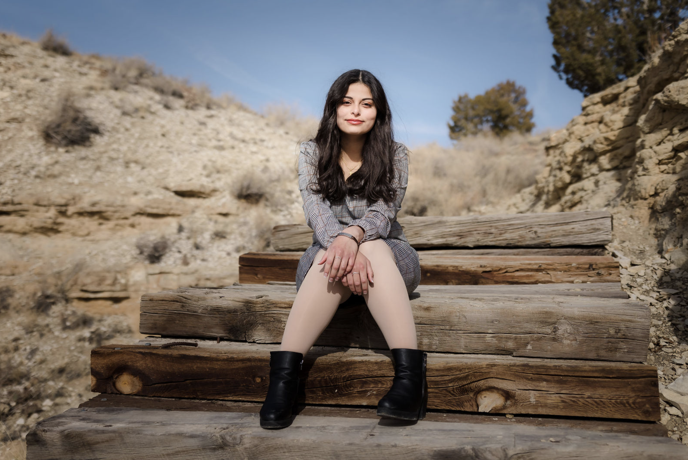 Young woman with dark hair sitting on wooden steps outdoors in a desert landscape with clear blue sky.