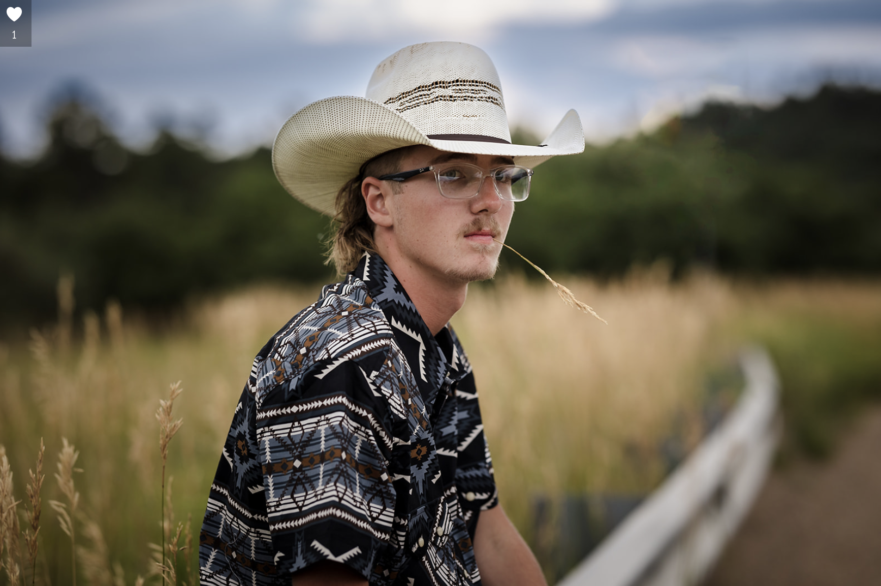 Young man with long hair wearing a cowboy hat, glasses, and a patterned shirt, sitting outdoors in a rural area with tall grass and trees in the background.