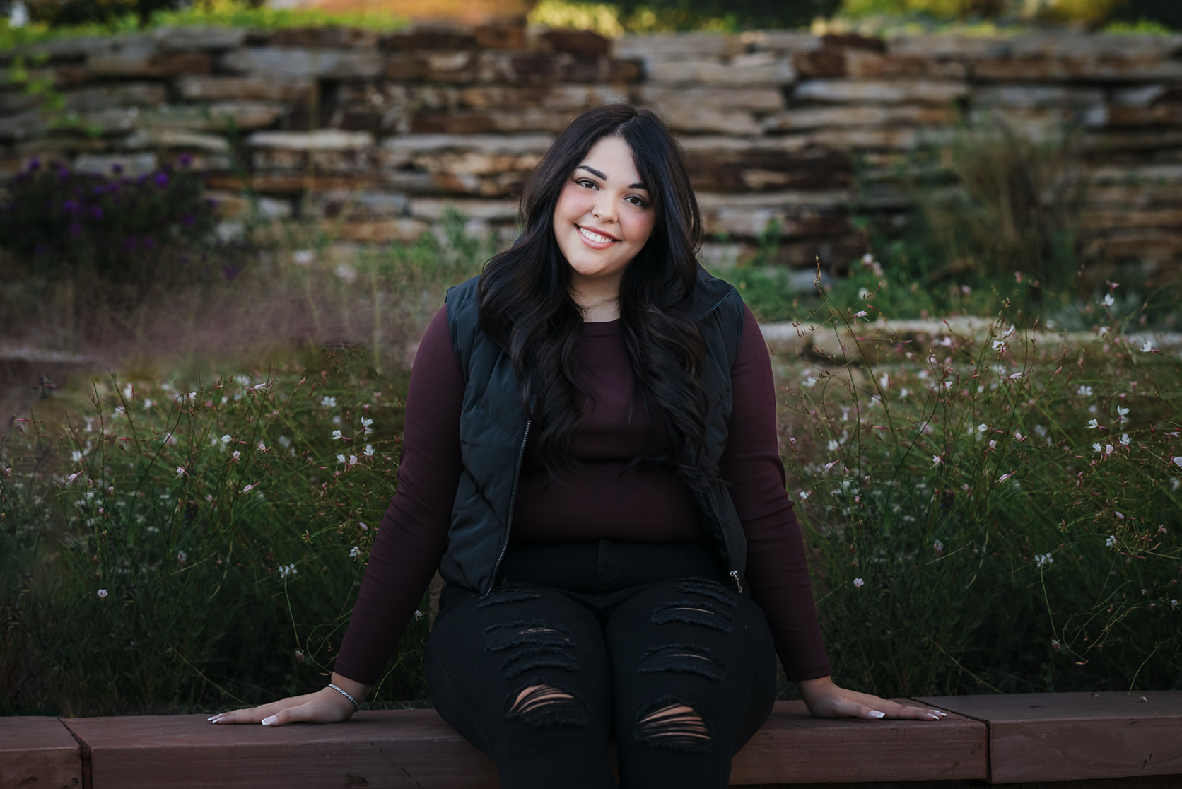 A young woman with long dark hair, wearing a black vest and ripped black jeans, sitting on a wooden bench outdoors surrounded by greenery and small flowers, smiling at the camera.