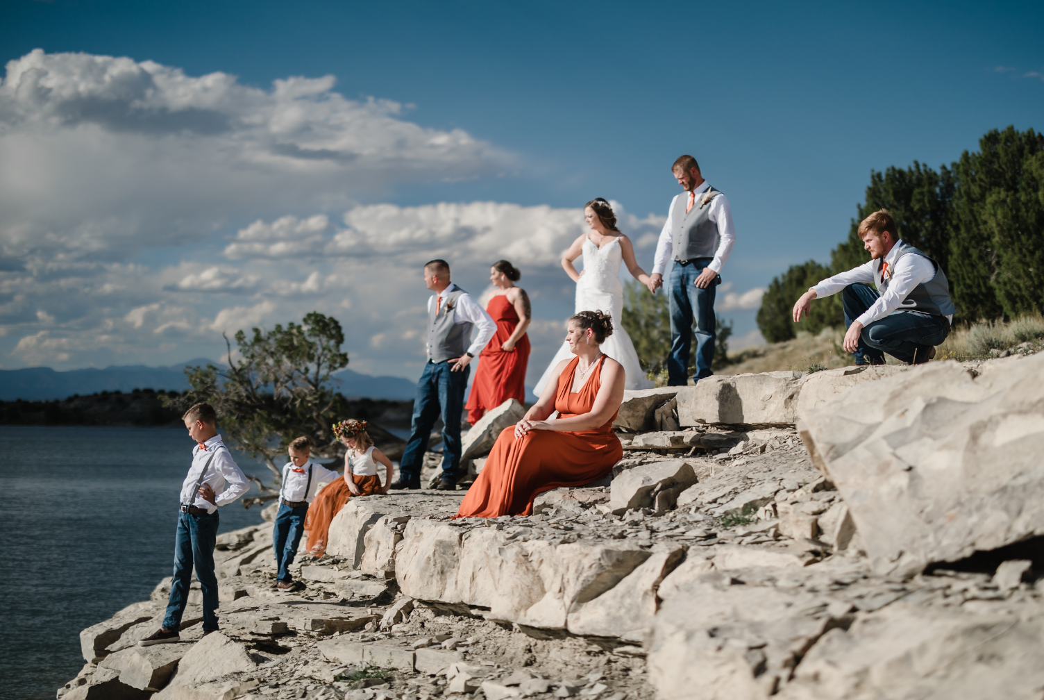 A woman and a young girl are smiling and hugging outdoors near a body of water during autumn, with colorful fall trees in the background.