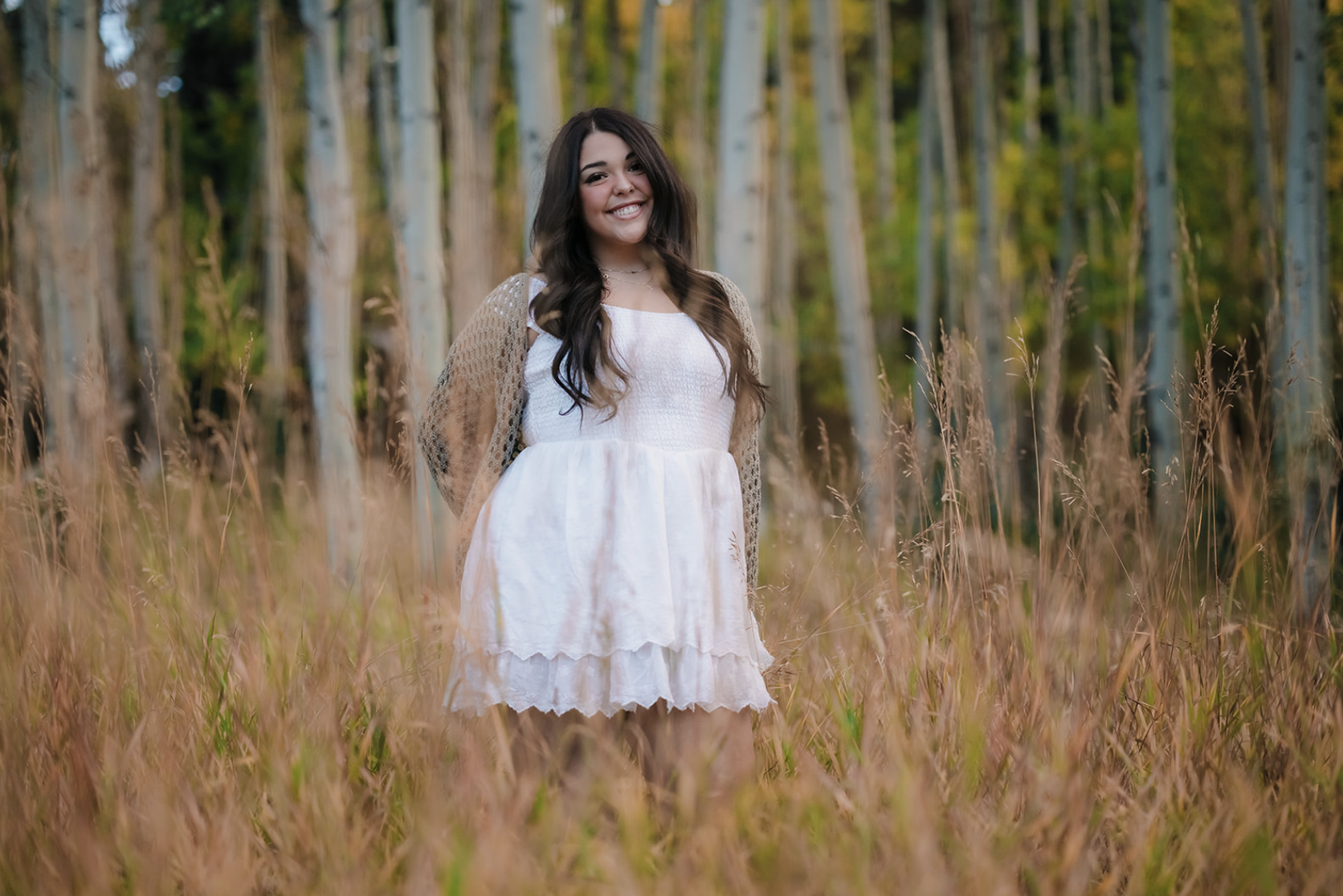 A woman in a white dress standing in a field of tall grass with a forest background, smiling.