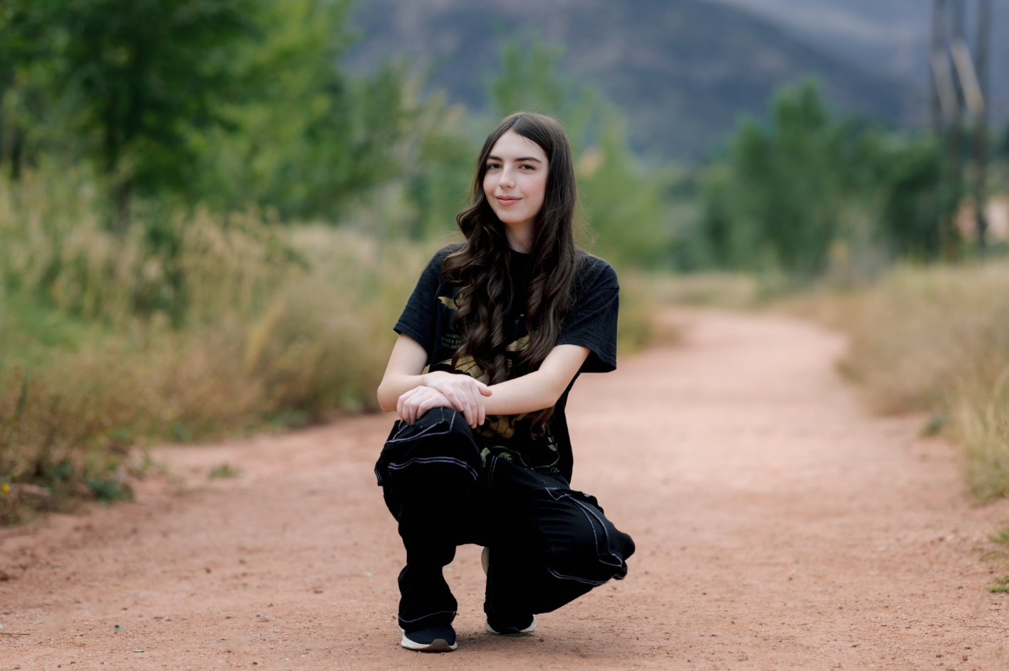A young woman with long dark hair kneeling on a dirt path in a natural outdoor setting with trees and greenery.