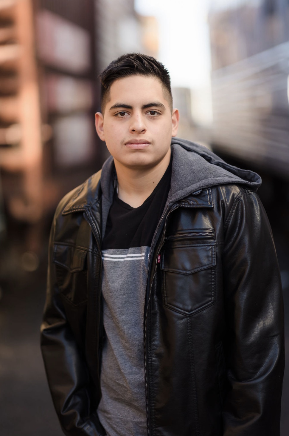Portrait of a young man with short dark hair, wearing a black leather jacket over a gray hoodie and a black shirt, standing outdoors in front of blurred background of buildings.