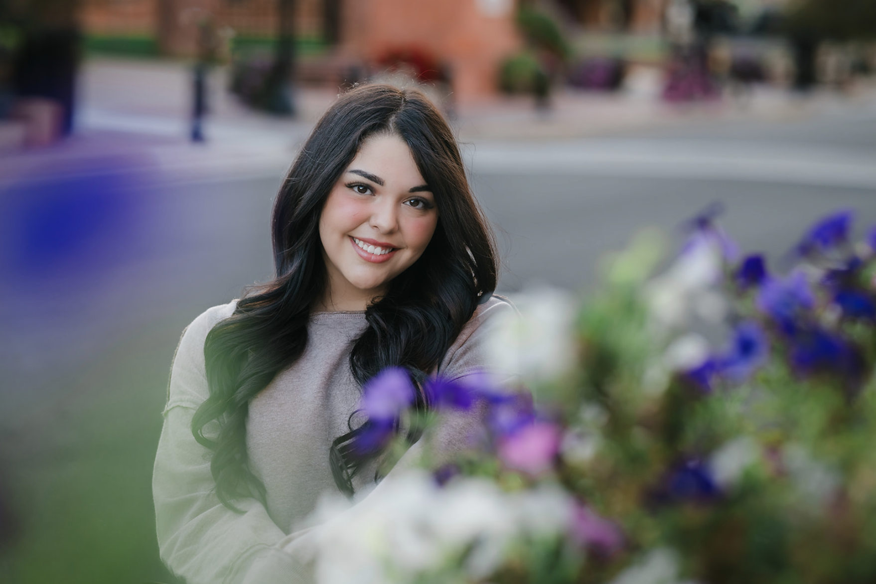 A young woman with long dark hair smiling outdoors next to colorful flowers with a blurred street background.