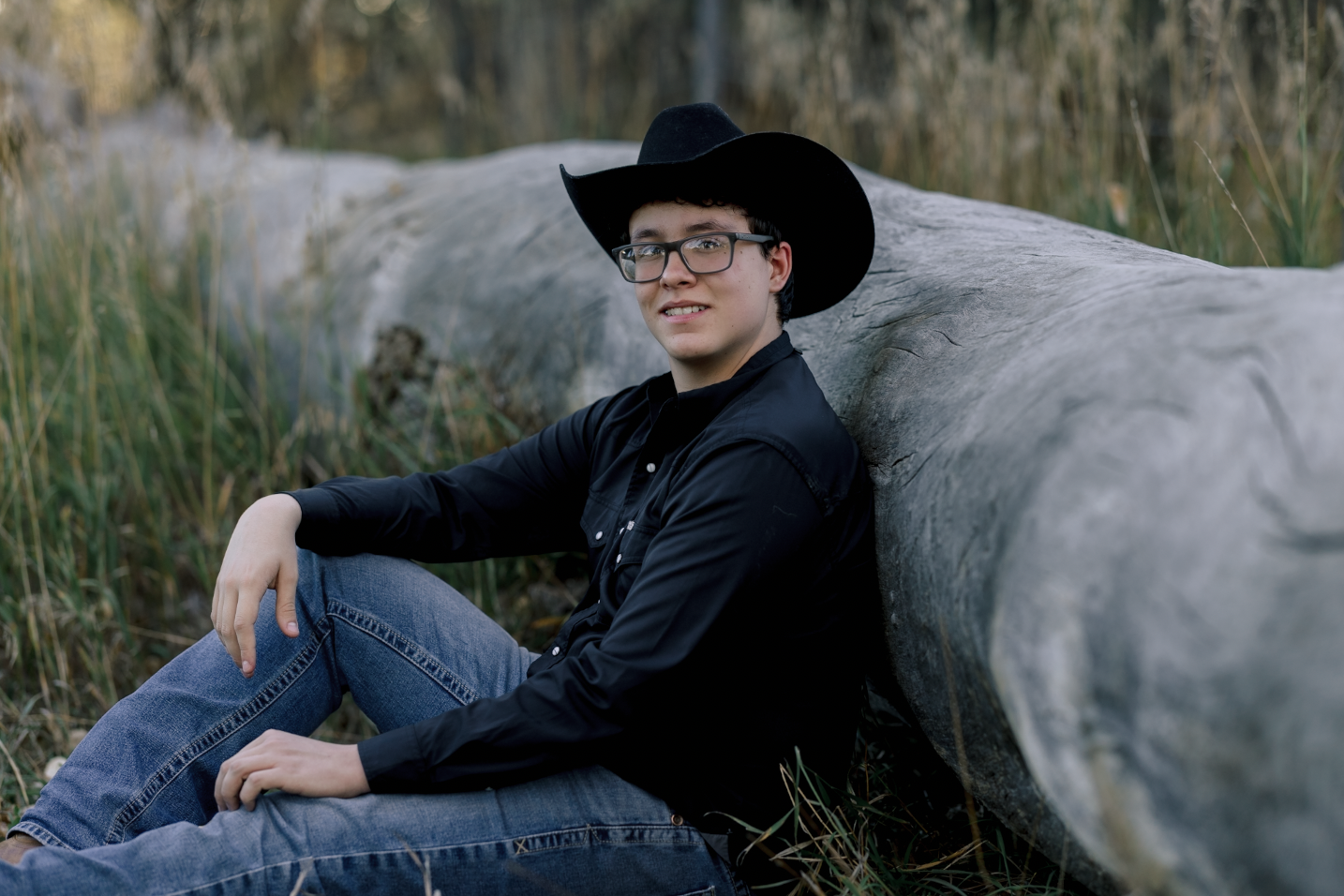 A young woman with curly brown hair, wearing a white dress, sitting on a fallen tree trunk in a lush green forest, smiling at the camera.