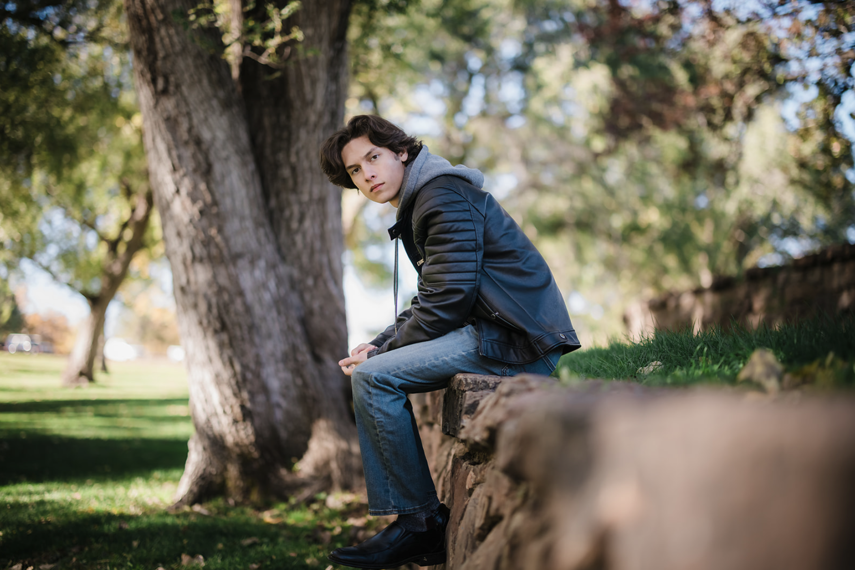 Young man sitting on a stone ledge in a park, wearing a leather jacket and jeans, with trees and greenery in the background.