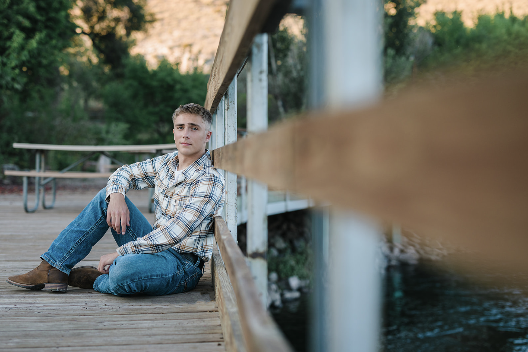 A young man sitting on a wooden deck next to a body of water, leaning against a railing, with trees in the background.
