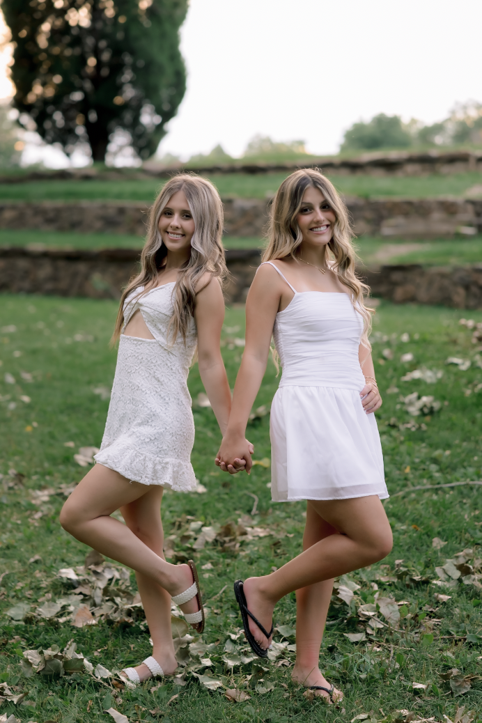 Two young women in white dresses holding hands and smiling, standing outdoors on grass surrounded by fallen leaves, with a tree and stepped terrain in the background.