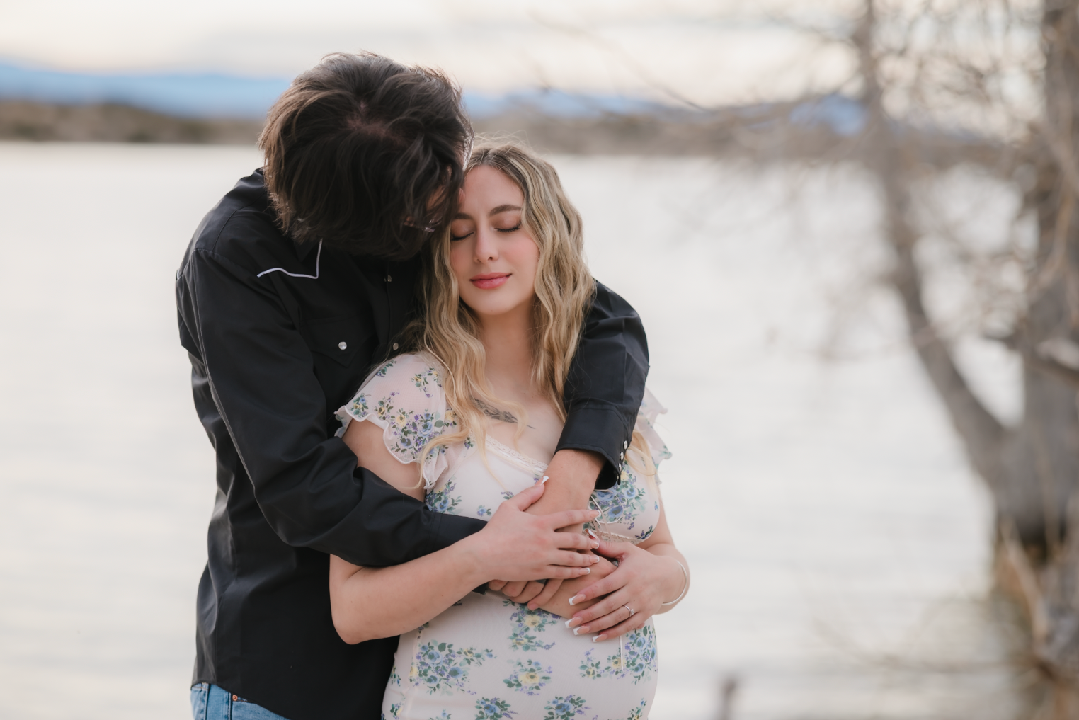A young woman with dark brown wavy hair and blue eyes smiling outdoors in a park during daytime, wearing a white sweater and layered gold necklaces.