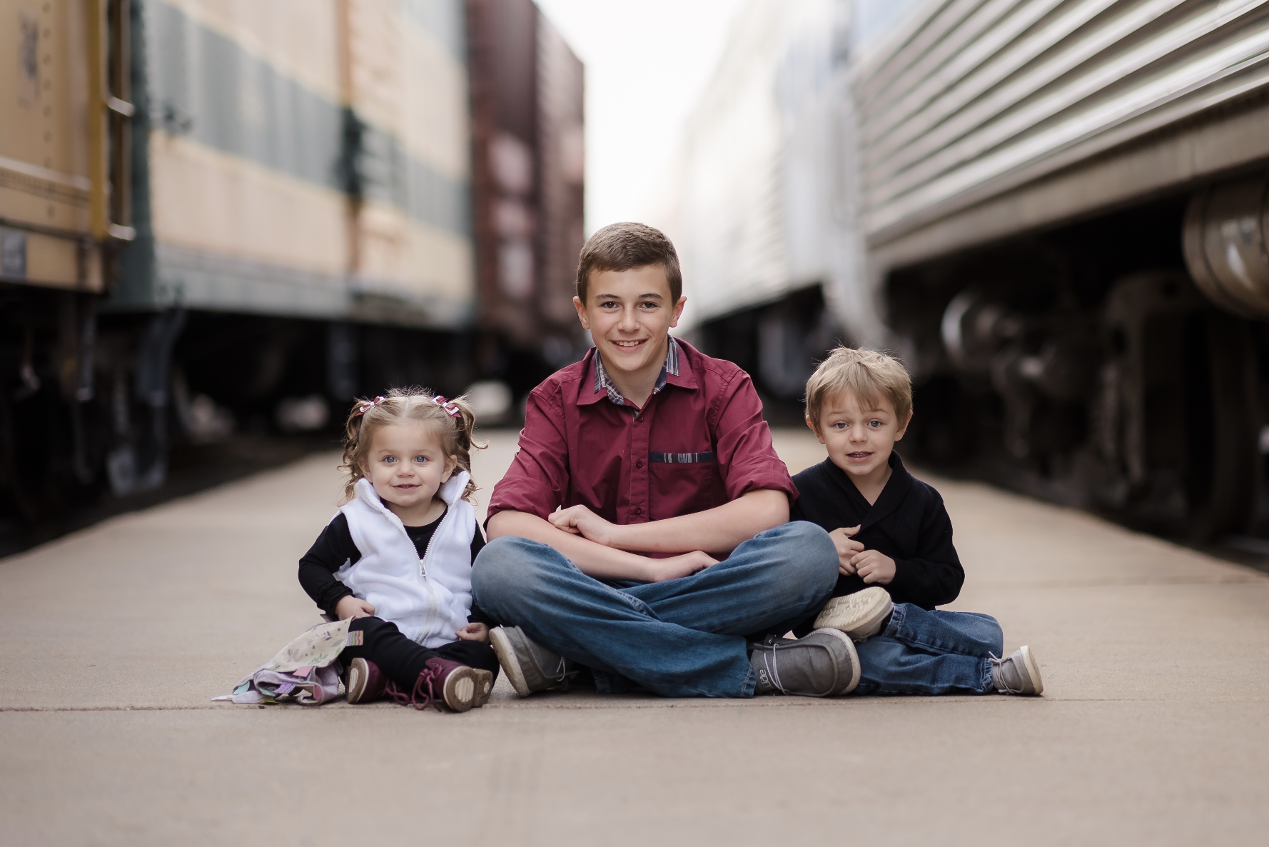 A mature couple stands close to each other by a lake with autumn trees in the background. The woman has gray hair and is wearing a brown sweater with hoop earrings and a necklace. The man has gray hair, glasses, and is wearing a plaid shirt. They are holding hands and looking at the camera.