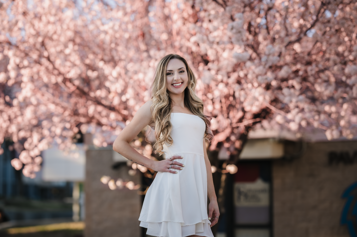 A young woman with long blonde hair in a white dress standing outdoors in front of a blooming pink cherry blossom tree, smiling with her hand on her hip.