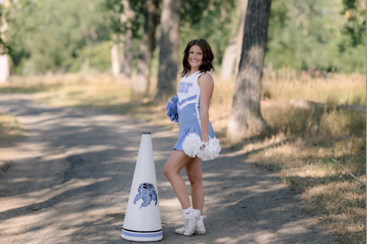 A smiling female cheerleader in a light blue cheerleading uniform stands on a dirt path, holding white pom-poms, with a cheerleader cone featuring a blue cartoon lion logo nearby, surrounded by trees and greenery.