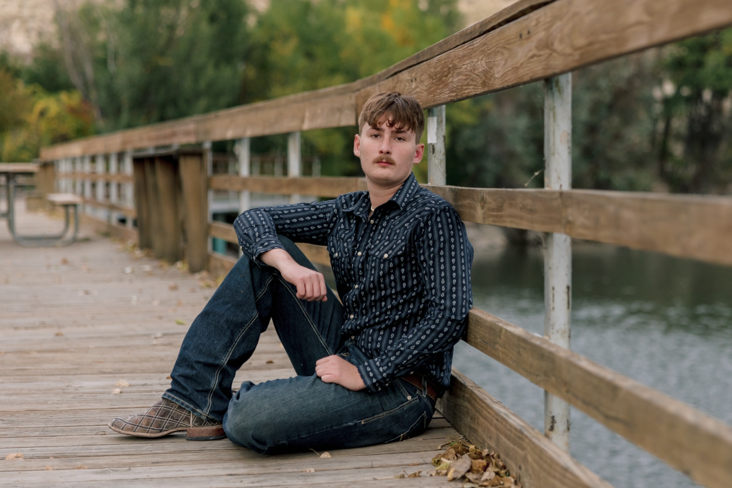 A young man with a mustache sitting on a wooden bridge by a river, wearing a dark patterned shirt and jeans, during autumn.