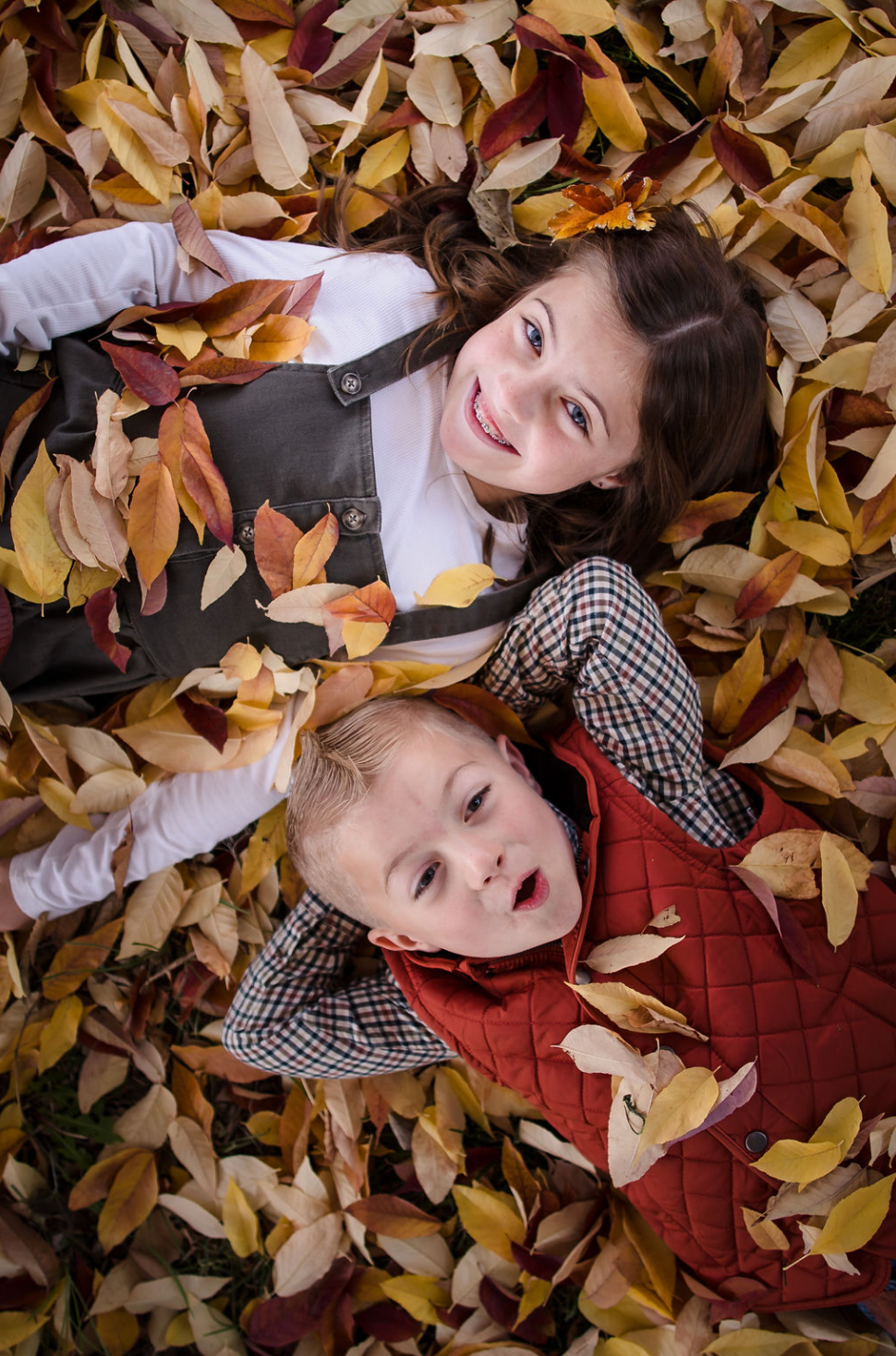 A woman and a young girl are smiling and hugging outdoors near a body of water during autumn, with colorful fall trees in the background.