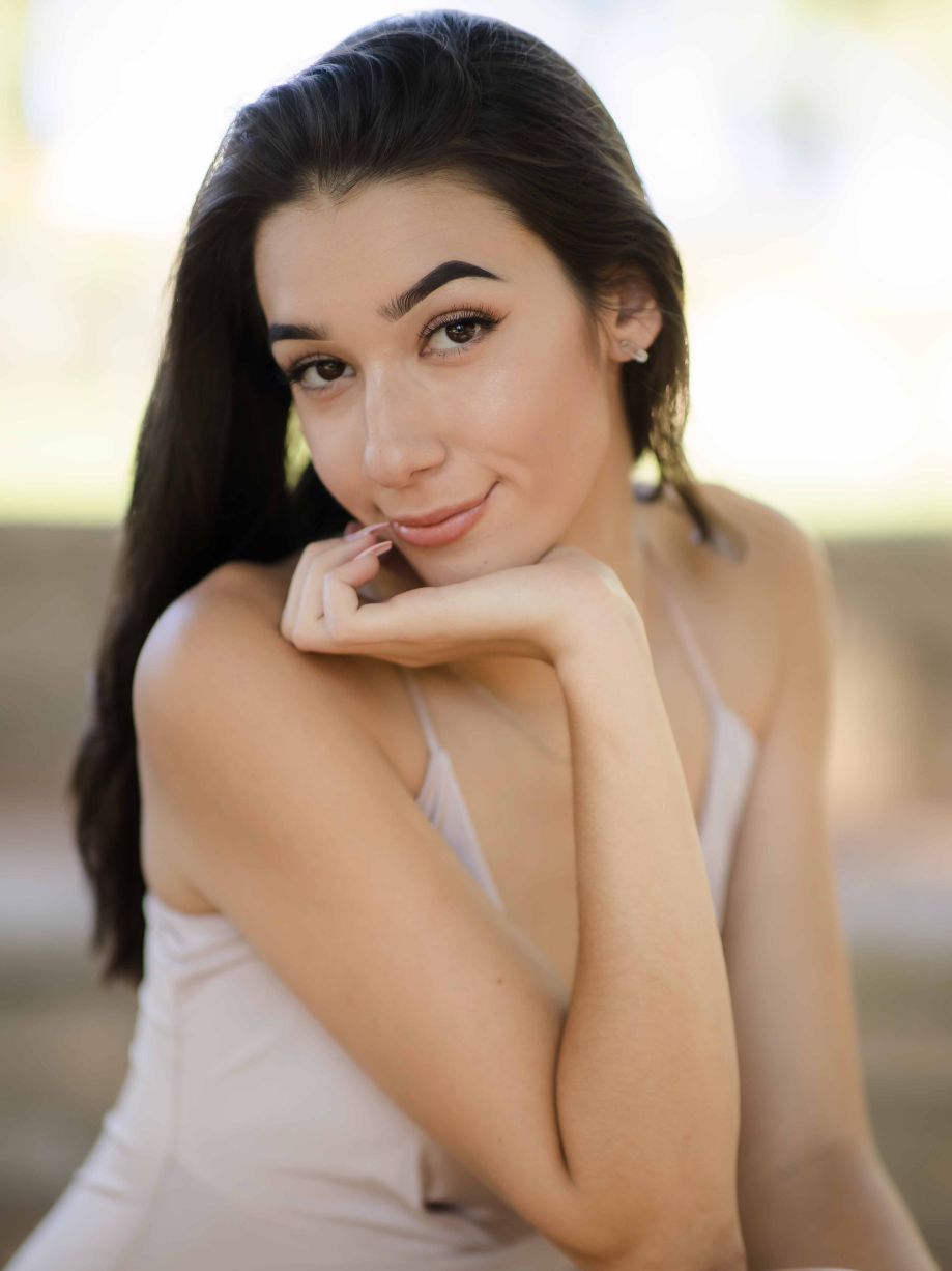 Close-up of a smiling young woman with long dark hair, wearing a light-colored top, posing with her chin resting on her hand against a blurred background.