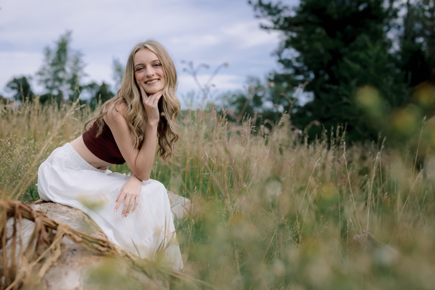 A smiling young woman with long blonde hair, wearing a brown crop top and a white skirt, sitting on a fallen log in a grassy field with trees in the background.