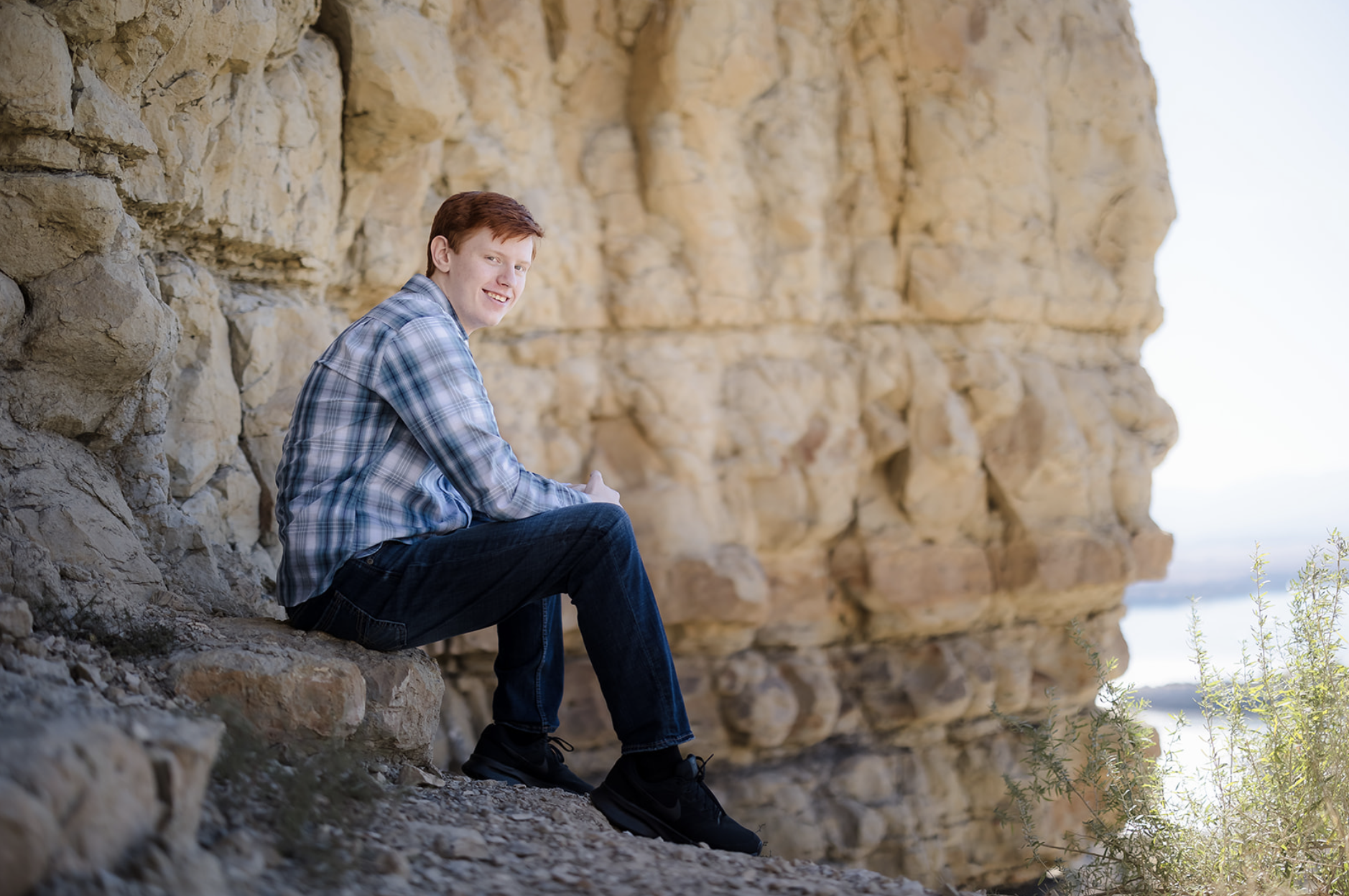 A young woman with dark brown wavy hair and blue eyes smiling outdoors in a park during daytime, wearing a white sweater and layered gold necklaces.