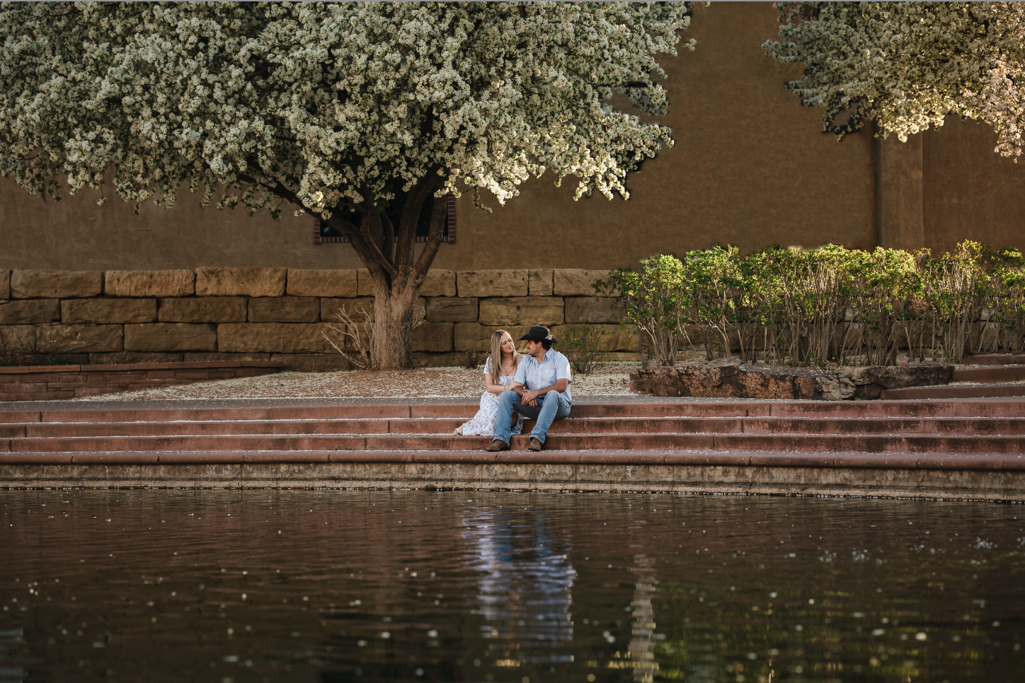 A young girl in a yellow dress with ruffled sleeves and black buttons sitting on a person's shoulder outdoors, smiling and looking at someone out of frame, with trees and greenery in the background.
