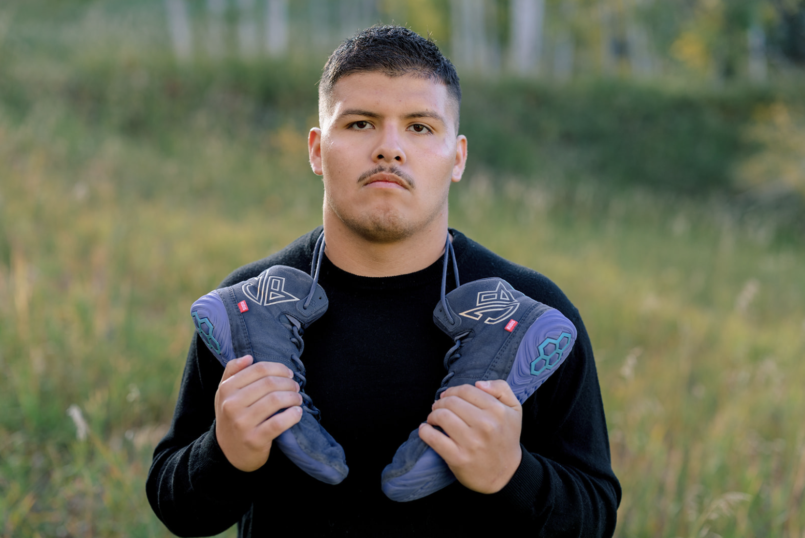 A young man outdoors holding a pair of black and purple sports shoes around his neck, with a neutral expression, standing in a grassy area with trees in the background.