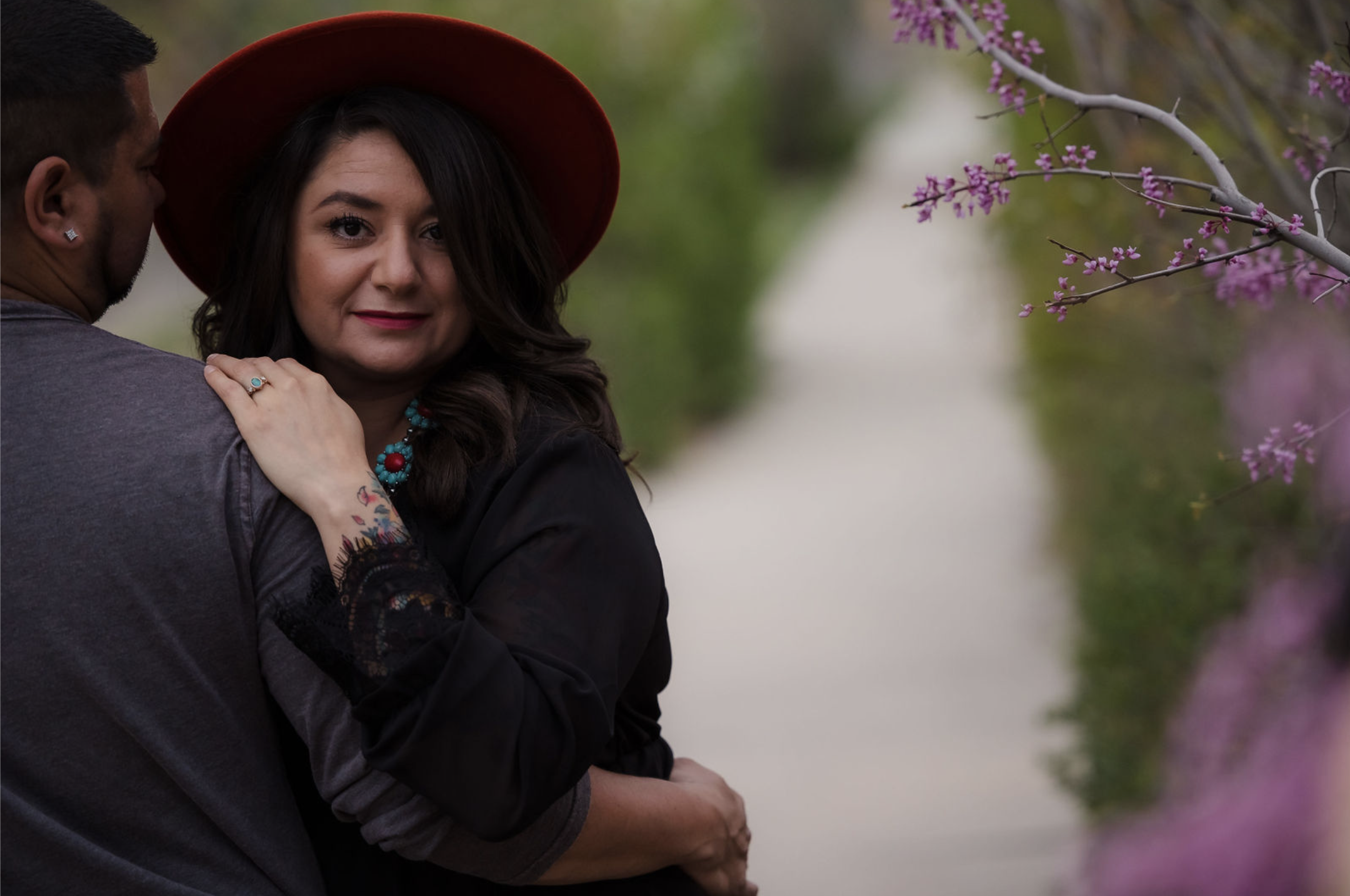 A young man with tousled blond hair and a serious expression, wearing a light gray T-shirt with buttons, standing outdoors in front of a calm body of water and a blurred green landscape.