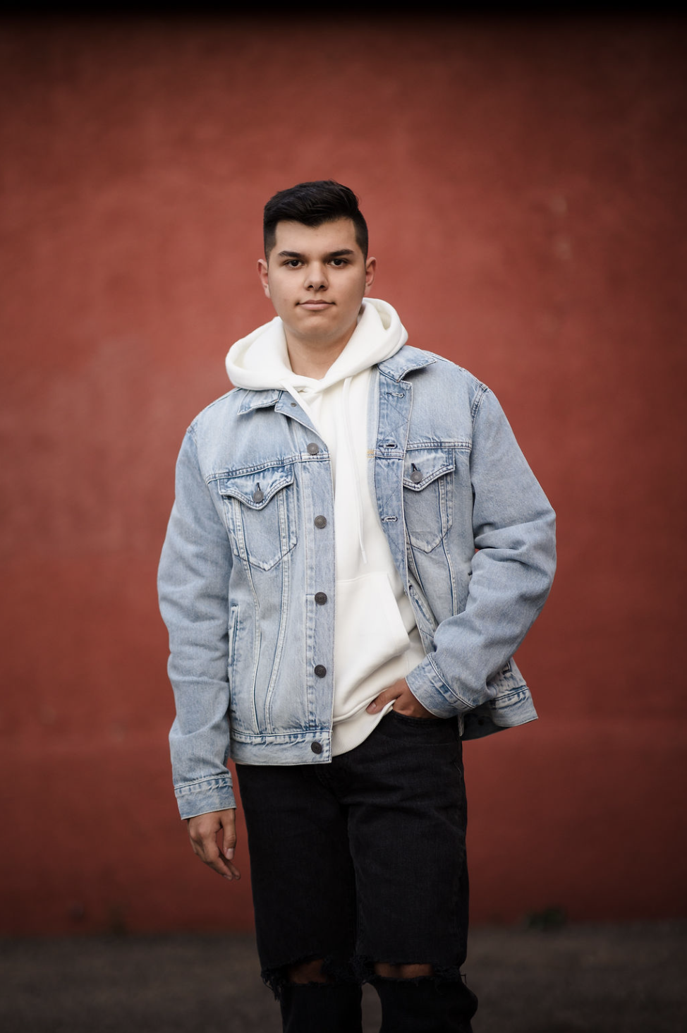 A young man wearing a denim jacket, white hoodie, and black ripped jeans, standing against a red wall.