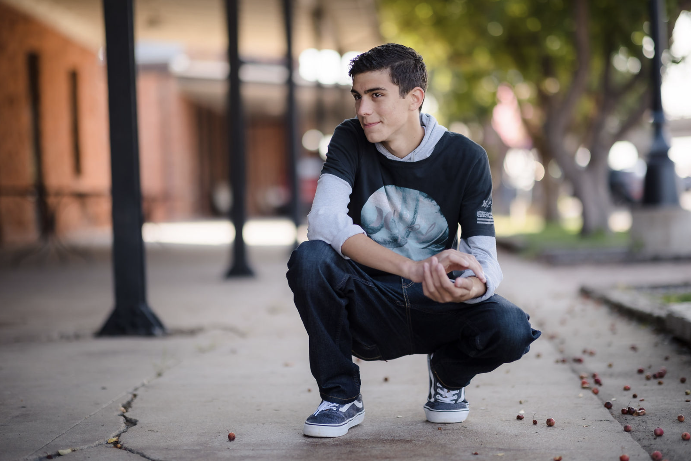 A young man squatting on a city sidewalk with a playful expression, surrounded by small red berries scattered on the ground, and trees and buildings in the background.