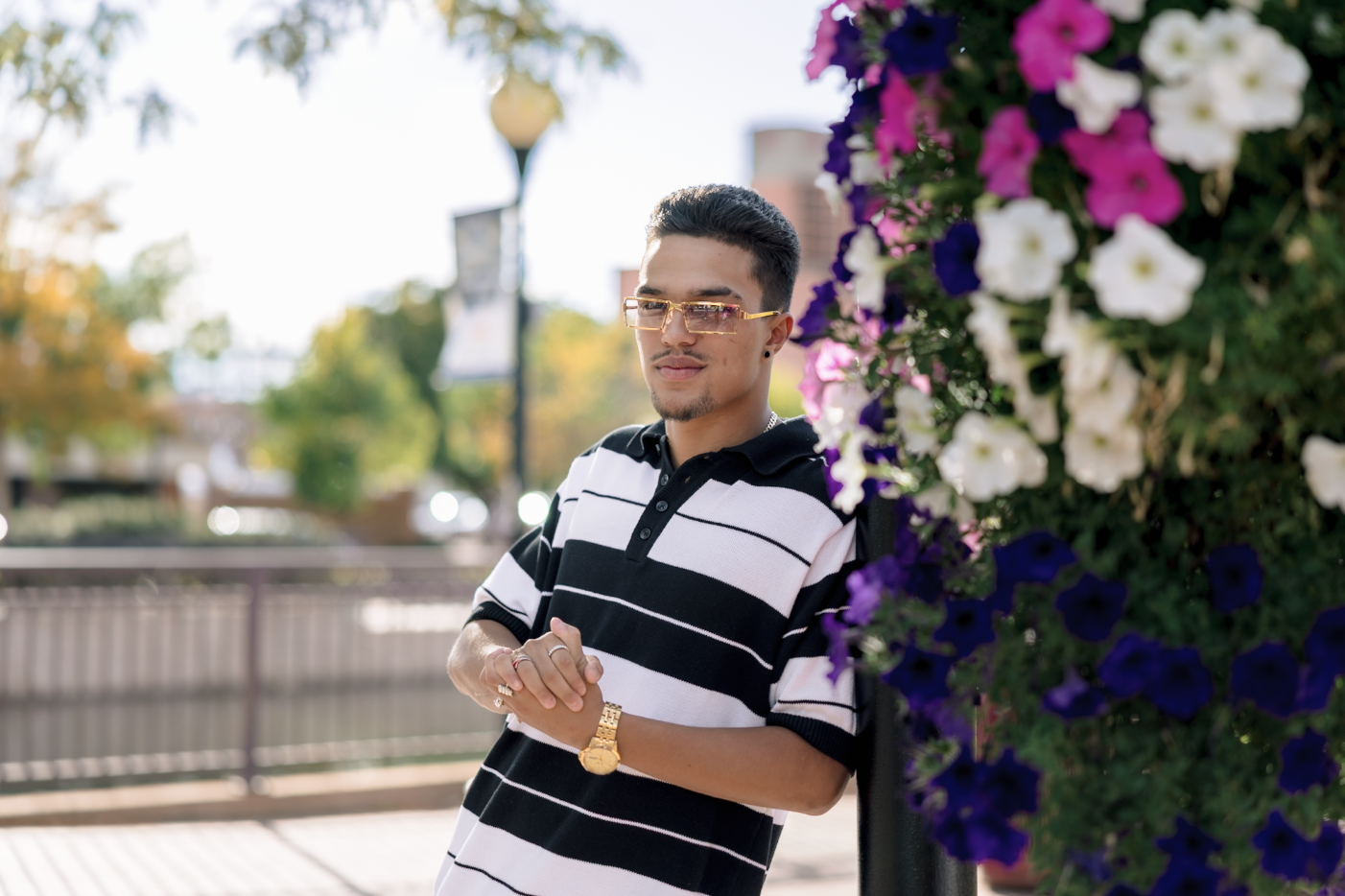 A young woman with dark brown wavy hair and blue eyes smiling outdoors in a park during daytime, wearing a white sweater and layered gold necklaces.