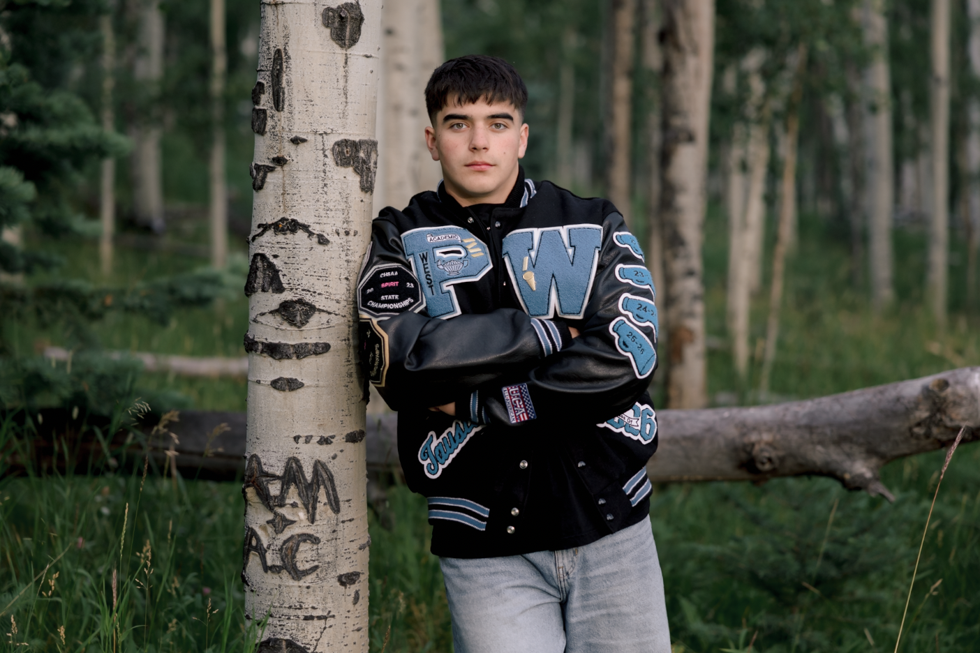 A young man with dark hair leaning against a tree in a forest, wearing a black varsity jacket with patches and light-colored pants.