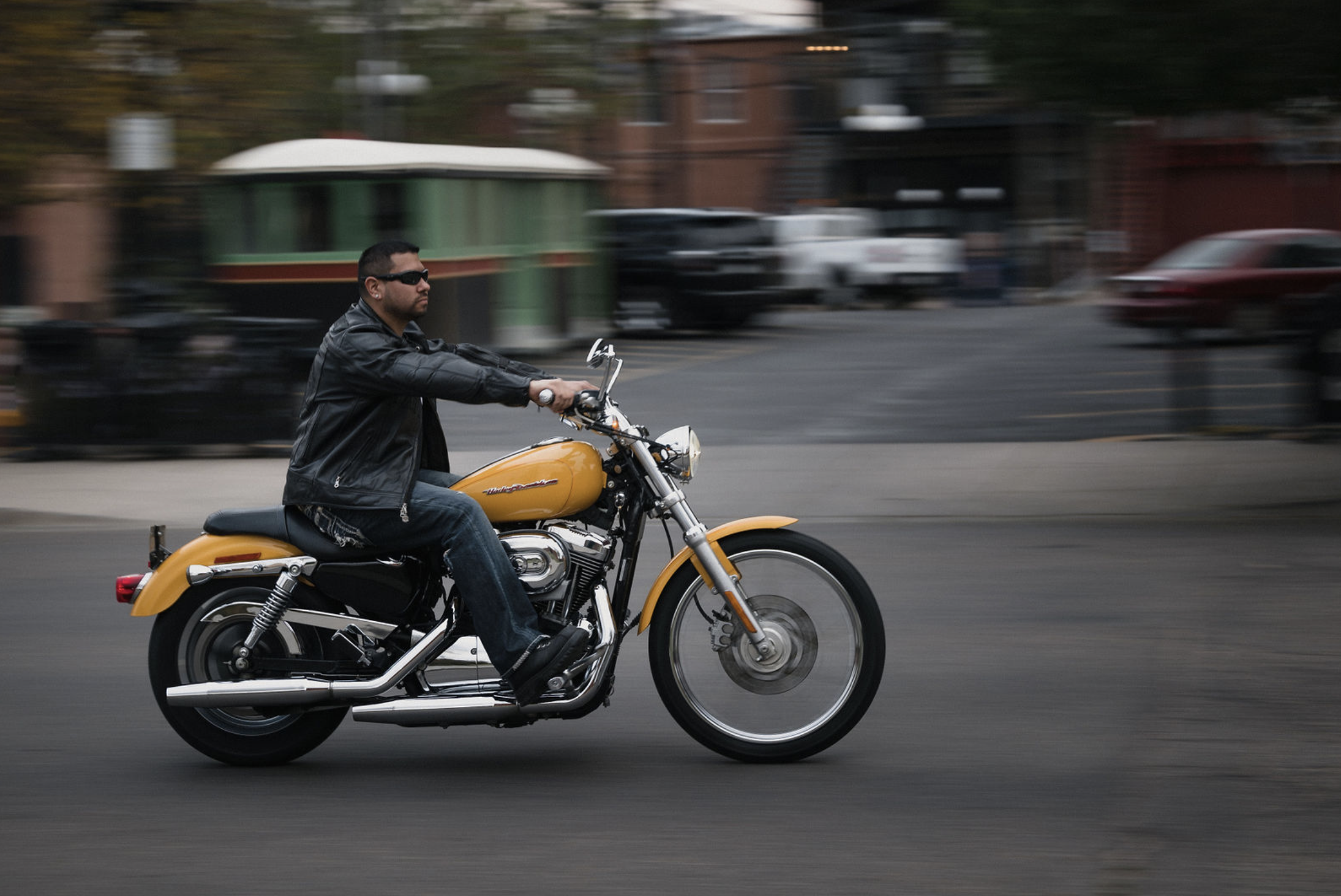 A man wearing sunglasses, a black leather jacket, and jeans riding a yellow Harley-Davidson motorcycle on a city street with blurred background.