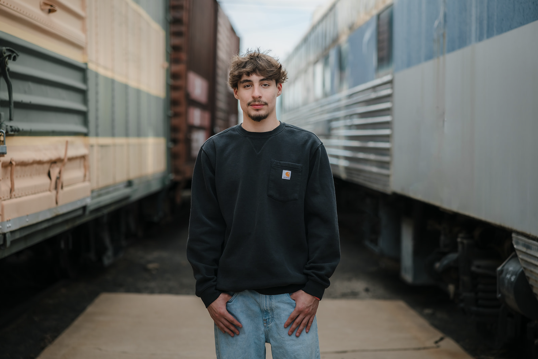 Young man standing between train cars, wearing a black sweatshirt and light blue jeans, outdoors.
