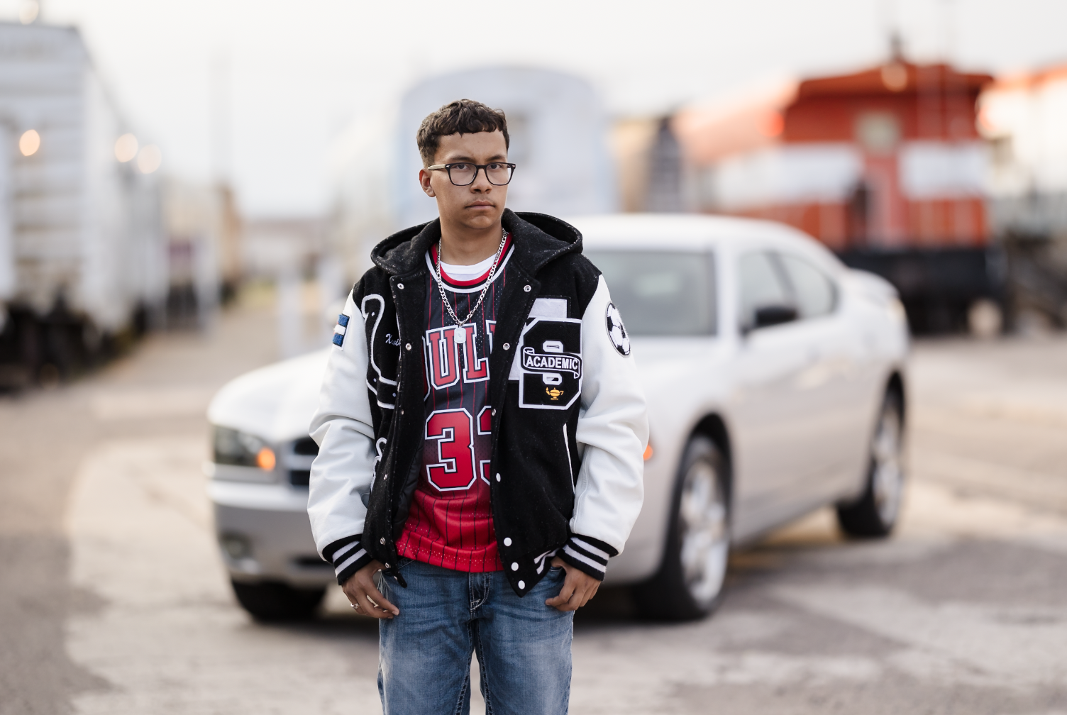 A young man wearing glasses and a black and white varsity jacket standing in front of a white car on an urban street.