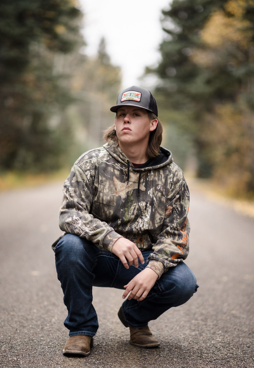 A young man wearing a camouflage hoodie, dark jeans, and brown boots squatting on a paved road surrounded by trees with autumn foliage, while wearing a black trucker hat with a colorful logo.