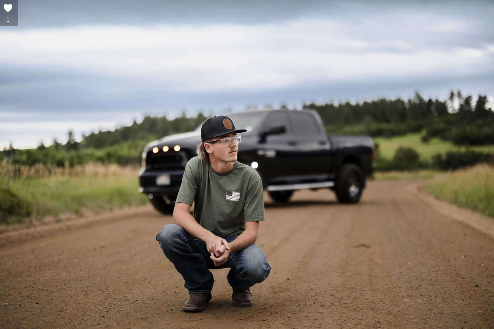 Young man crouching on a dirt road with a black truck behind him, surrounded by green fields and trees under a cloudy sky.