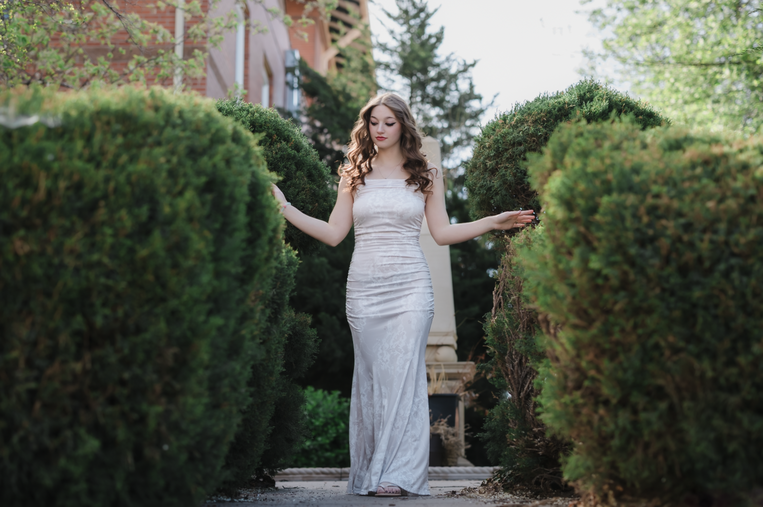A young woman with curly brown hair, wearing a white dress, sitting on a fallen tree trunk in a lush green forest, smiling at the camera.