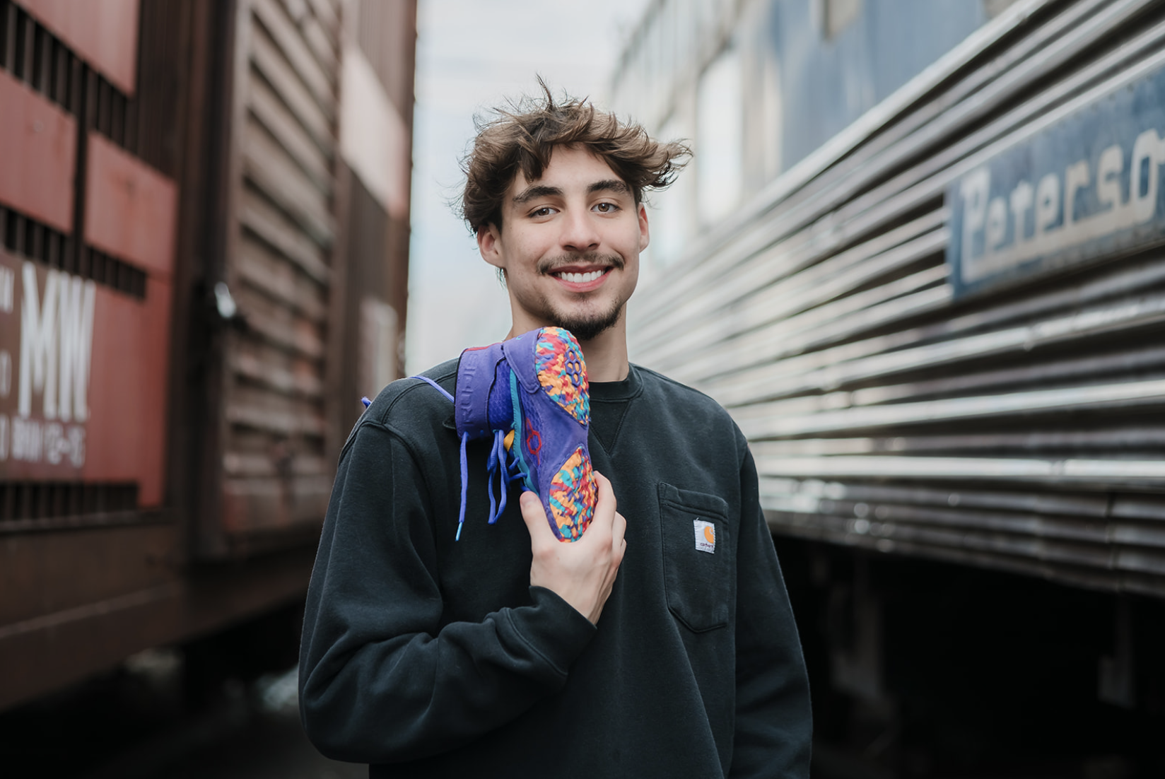 A young man with brown hair and a goatee smiling while carrying a colorful backpack on his shoulder, standing between train cars.
