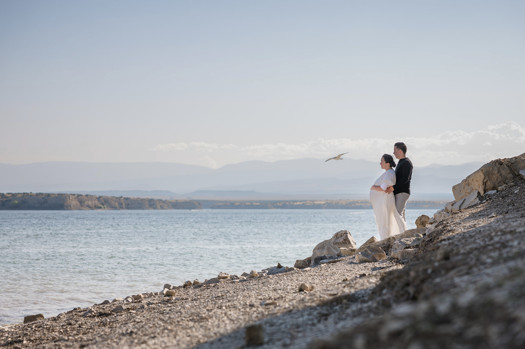 Two children playing near the edge of a lake during sunset, with trees in the background and the sun reflecting on the water.