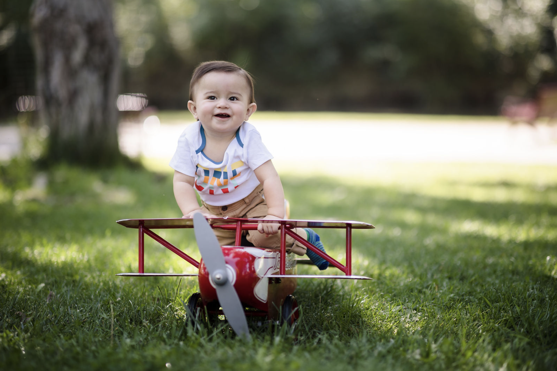 A young man with tousled blond hair and a serious expression, wearing a light gray T-shirt with buttons, standing outdoors in front of a calm body of water and a blurred green landscape.