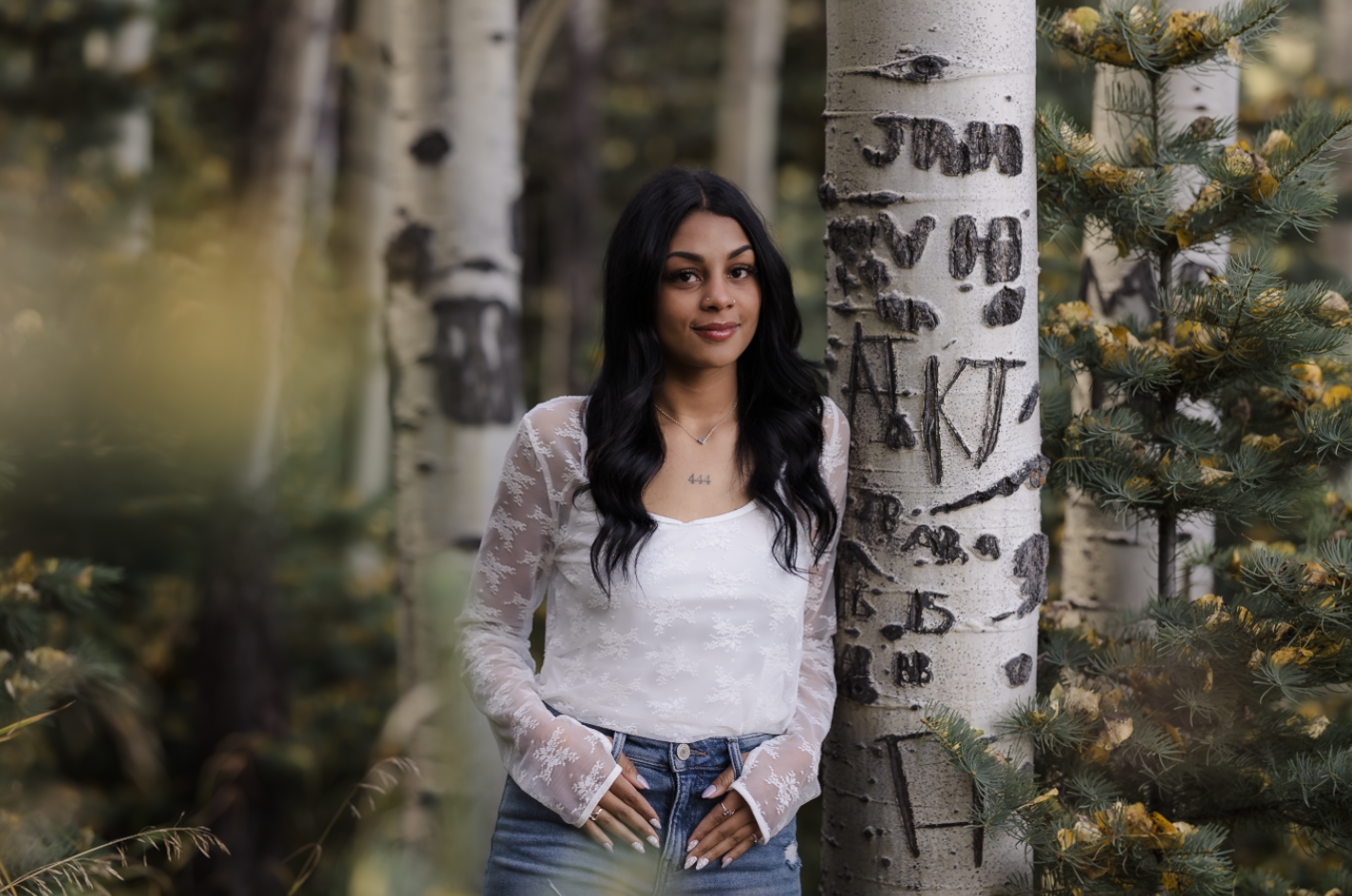 A woman with long black hair and a white lace blouse standing outdoors next to a birch tree, with green foliage and other trees in the background.