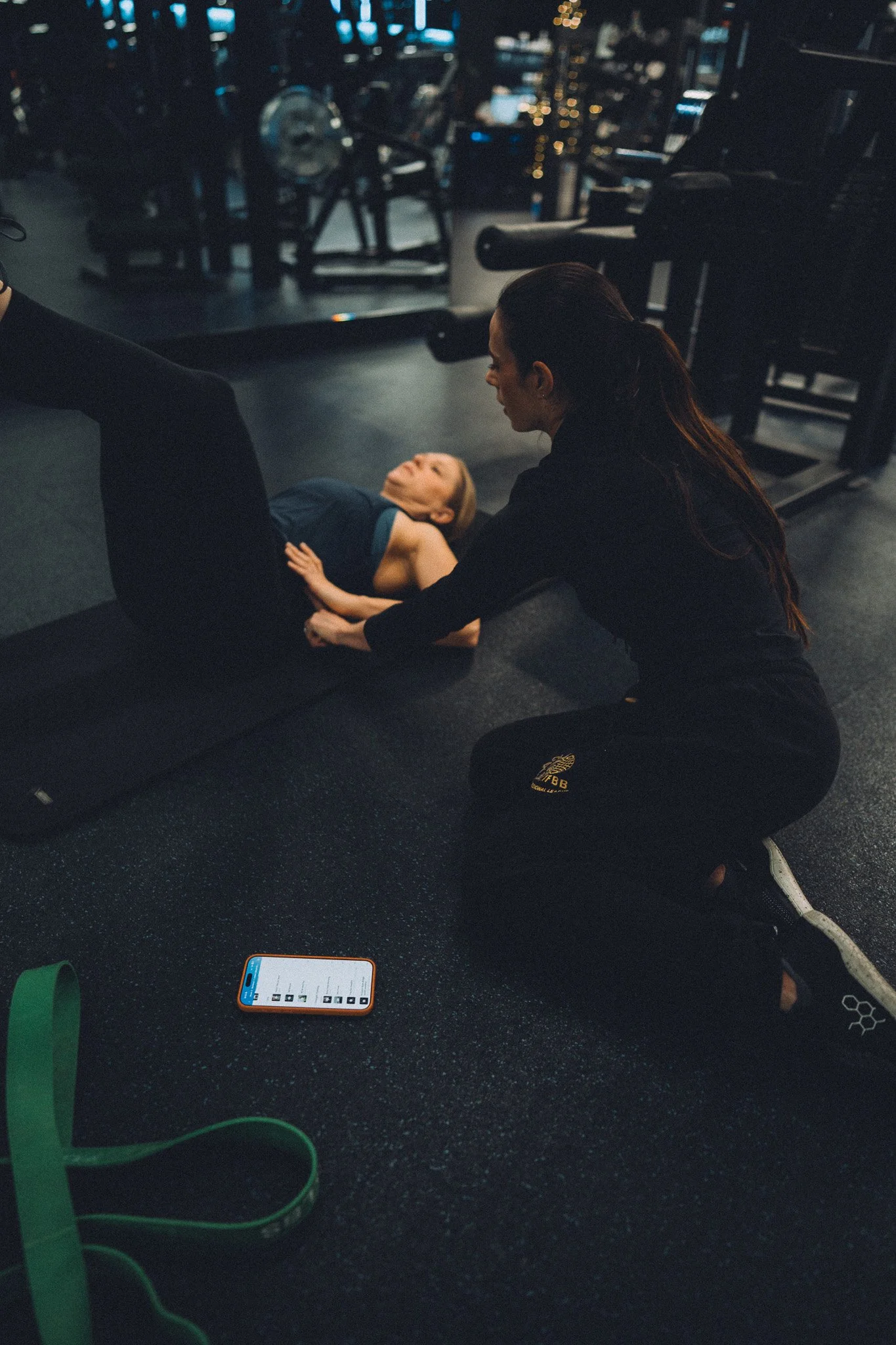 A woman with long dark hair kneeling on the floor helping a young girl do sit-ups in a gym. The girl is lying on her back with her legs bent and her head lifted. Gym equipment and a smartphone are visible in the background.